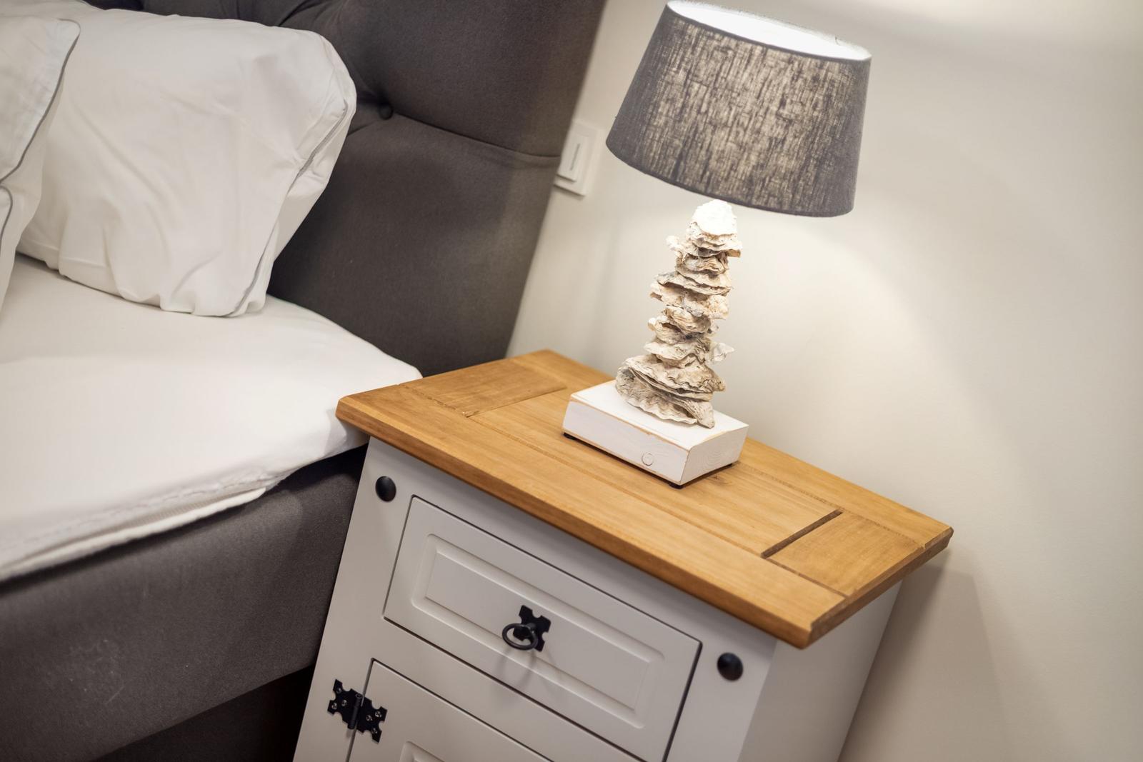 Nachttisch mit Holzoberfläche, Lampe und Bett im Hintergrund.