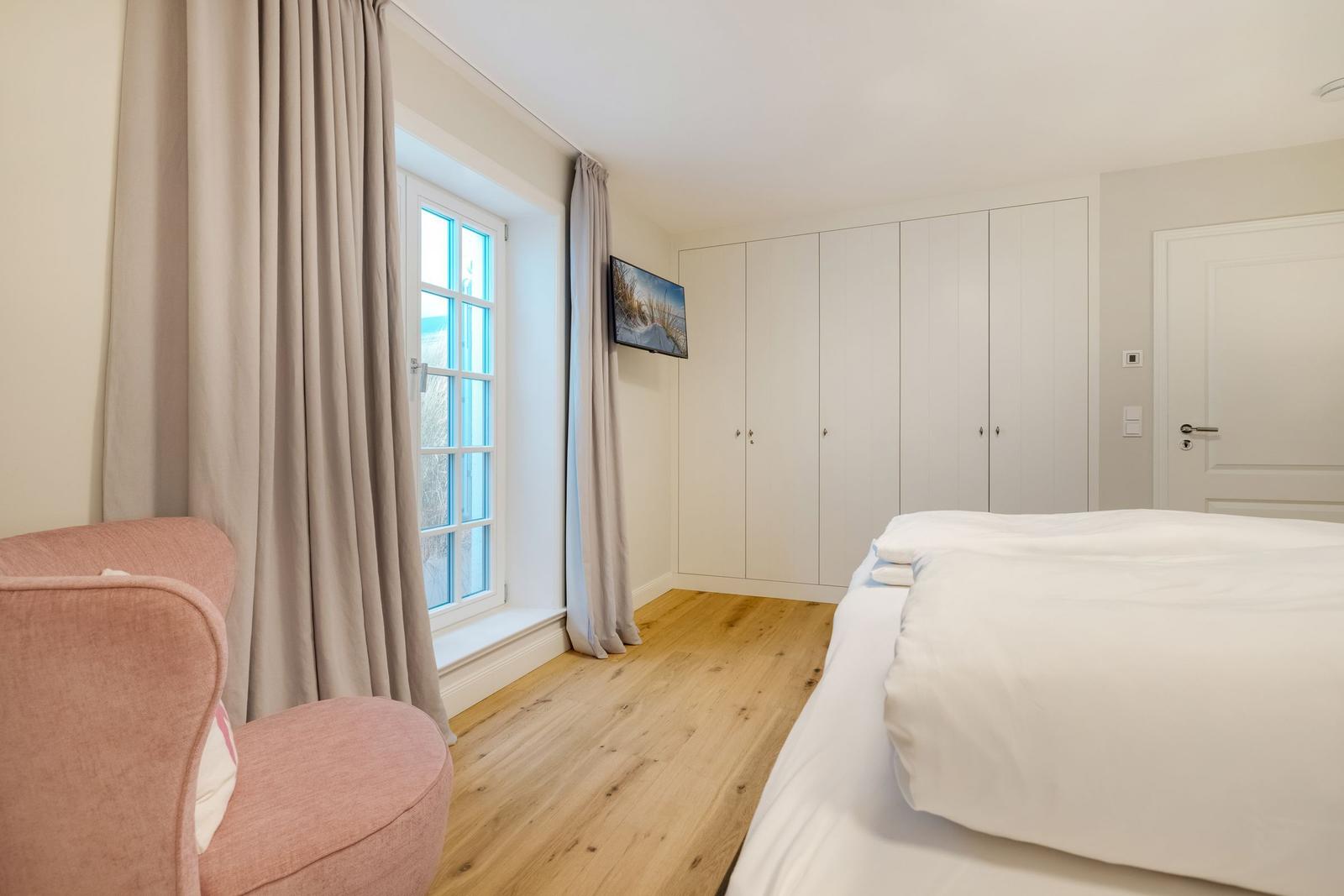Schlafzimmer mit Bett, rosa Sessel, Fenster und Kleiderschrank.