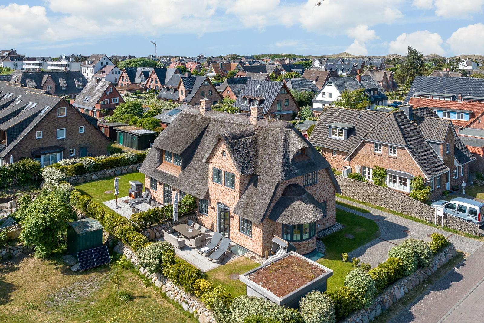 Haus mit Strohdach, Garten und Terrasse. Blick auf Wohnsiedlung und Hügel.