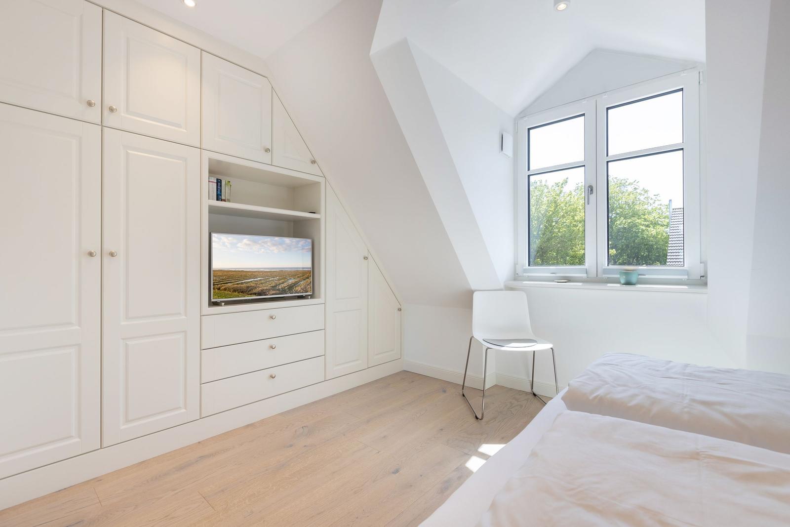 Schlafzimmer mit Dachschrägen, weißer Einbauschränke, Fernseher und Fenster mit Blick auf Bäume.