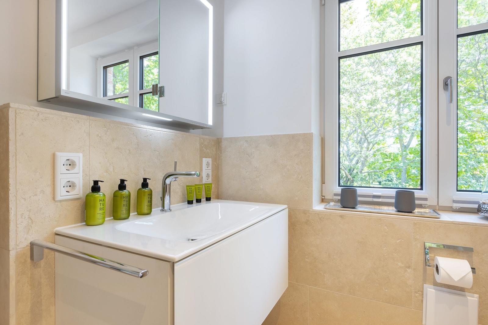 Badezimmer mit Waschbecken, Spiegel und Fenster mit Blick auf Bäume.