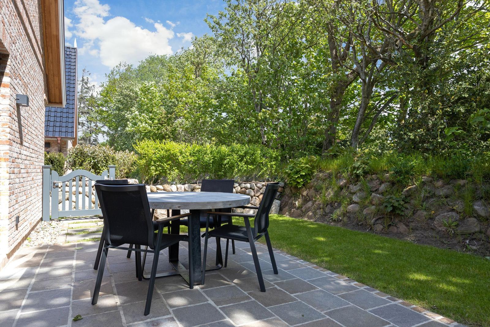 Terrasse mit Tisch und Stühlen neben Haus, Grünfläche und Bäume im Hintergrund.