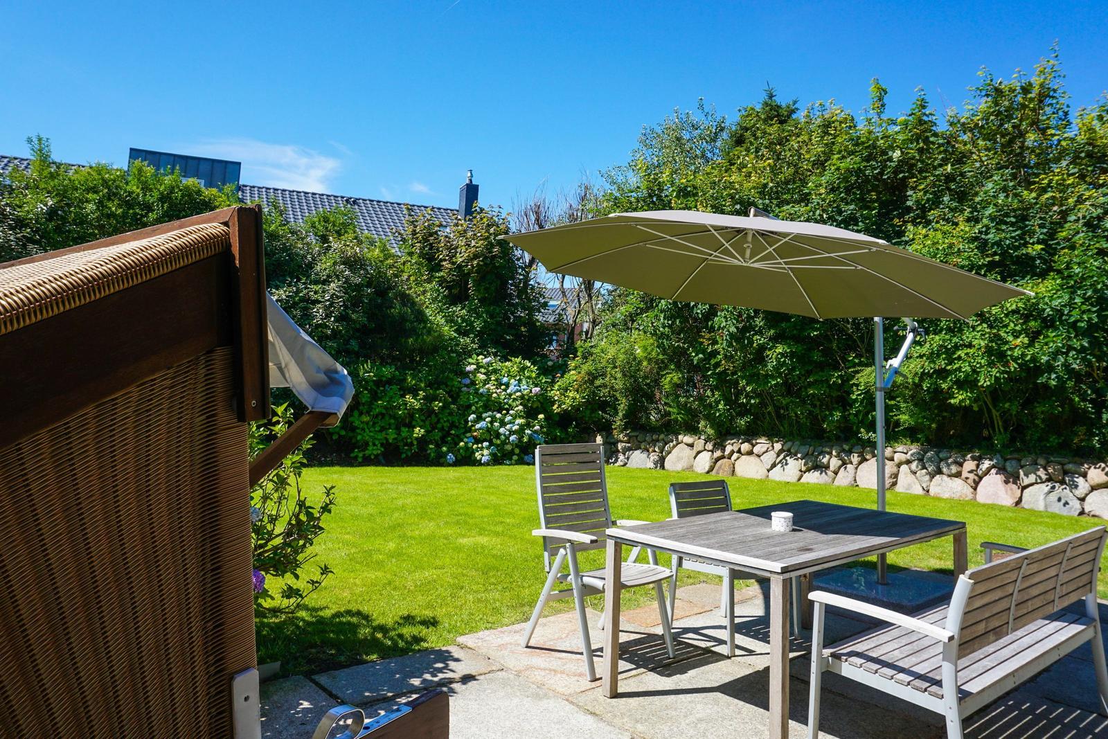 Terrace with table, chairs, and umbrella. Lawn and hedge in the background.