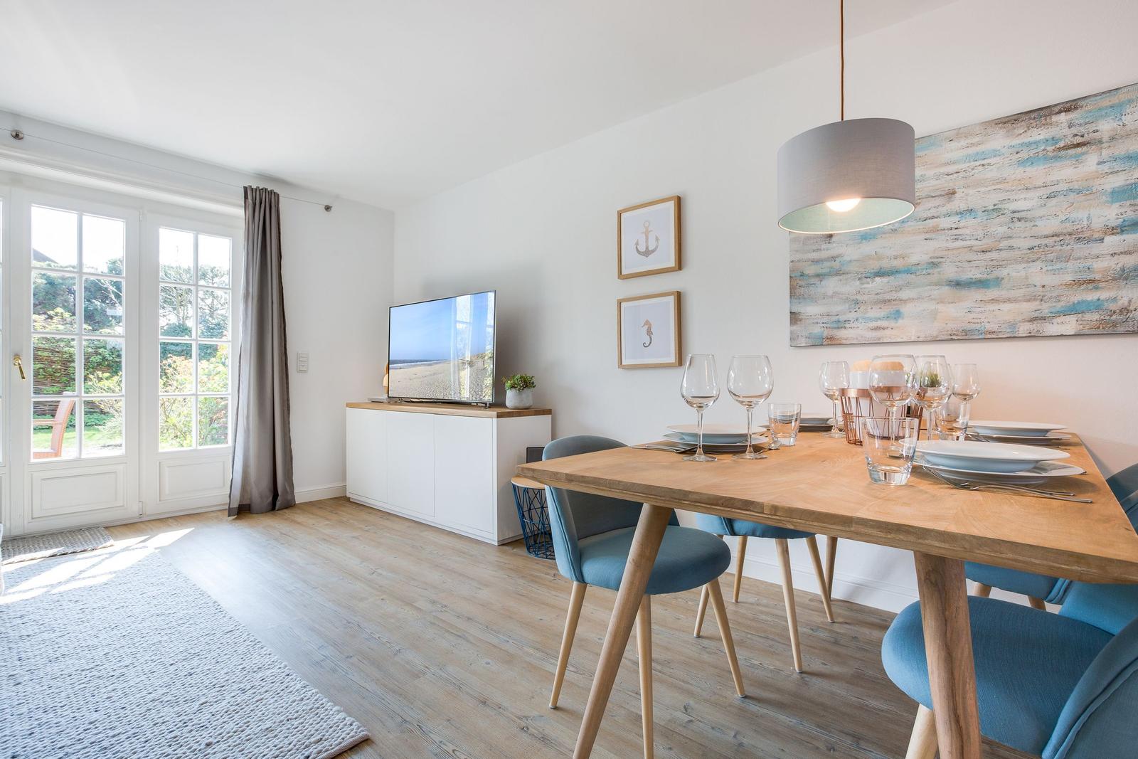 Essbereich mit Holztisch, blauen Stühlen und TV. Fenster zur Außenterrasse.