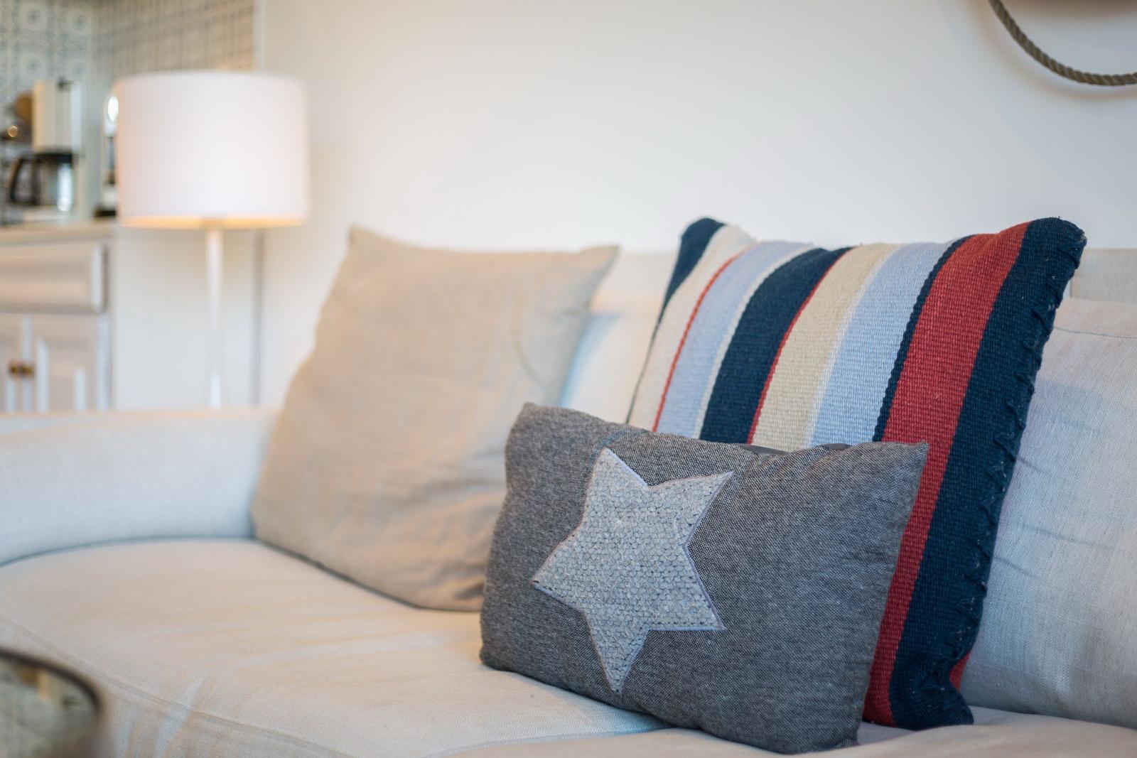 A sofa with striped and star-patterned cushions. Background: lamp and kitchen area.