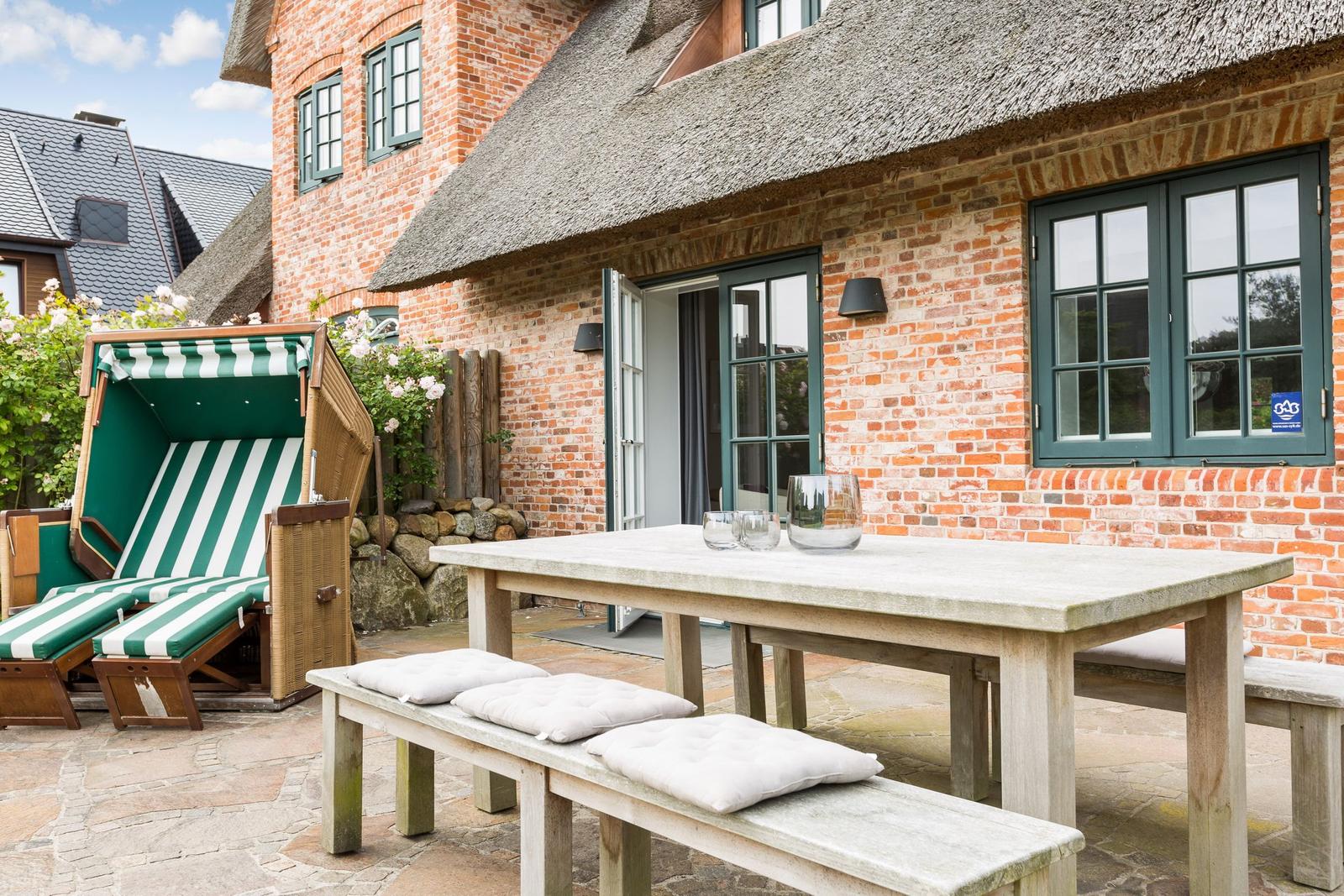 Außenbereich mit Holztisch, Bänken und Strandkörben vor einem Backsteinhaus mit Strohdach.