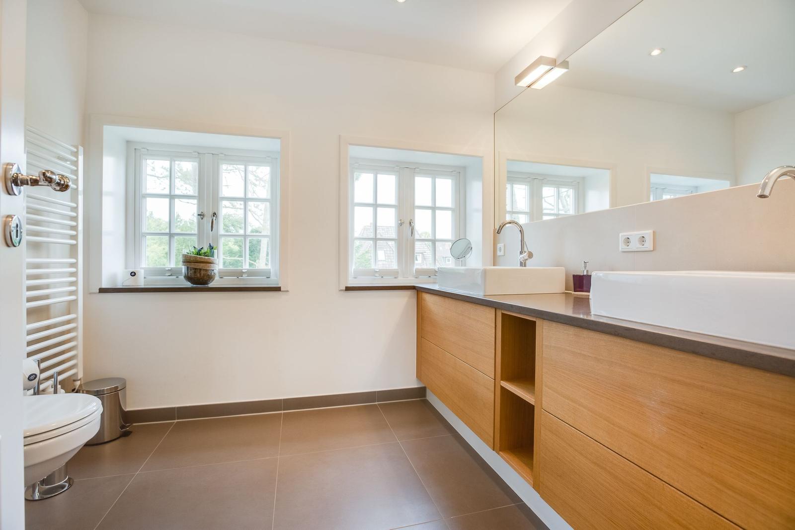 Badezimmer mit Holzschrank, zwei Fenstern und Spiegel.
