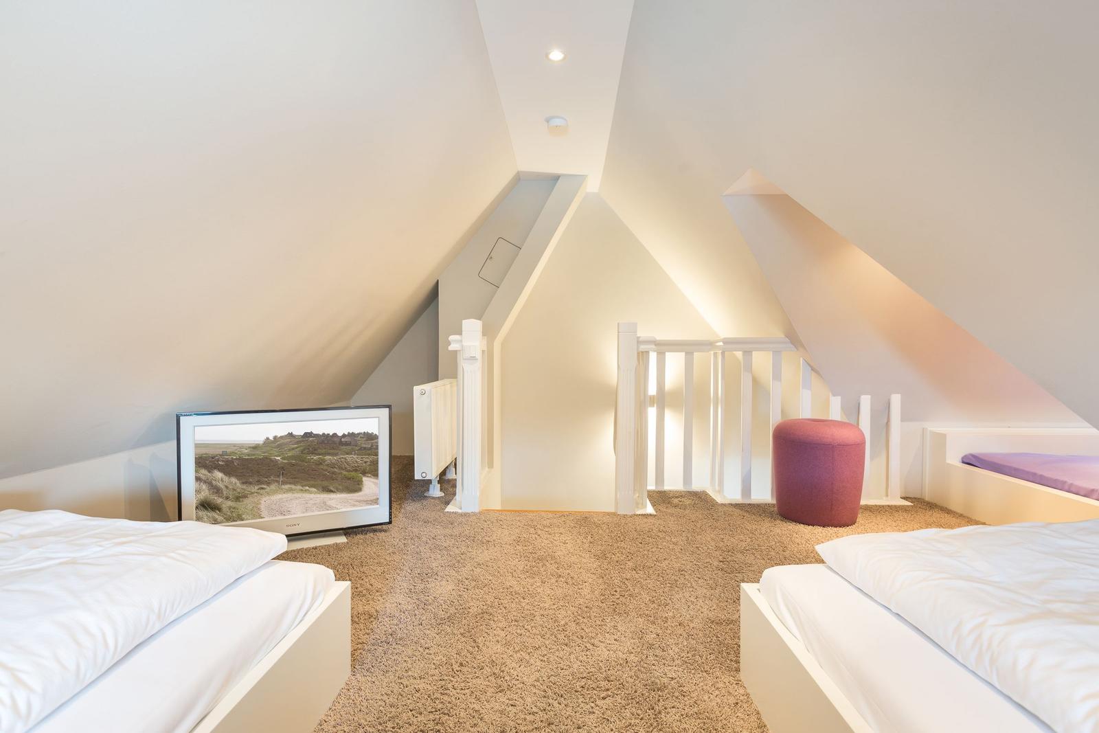 Zimmer mit zwei Betten, Fernseher und rotem Sitzkissen unter schrägen Wänden.