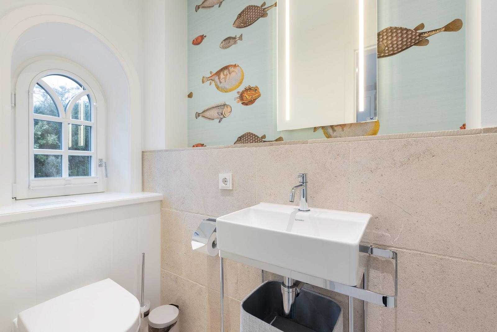 Badezimmer mit Fischmotivwand, Waschbecken und Toilette unter einem Bogenfenster.