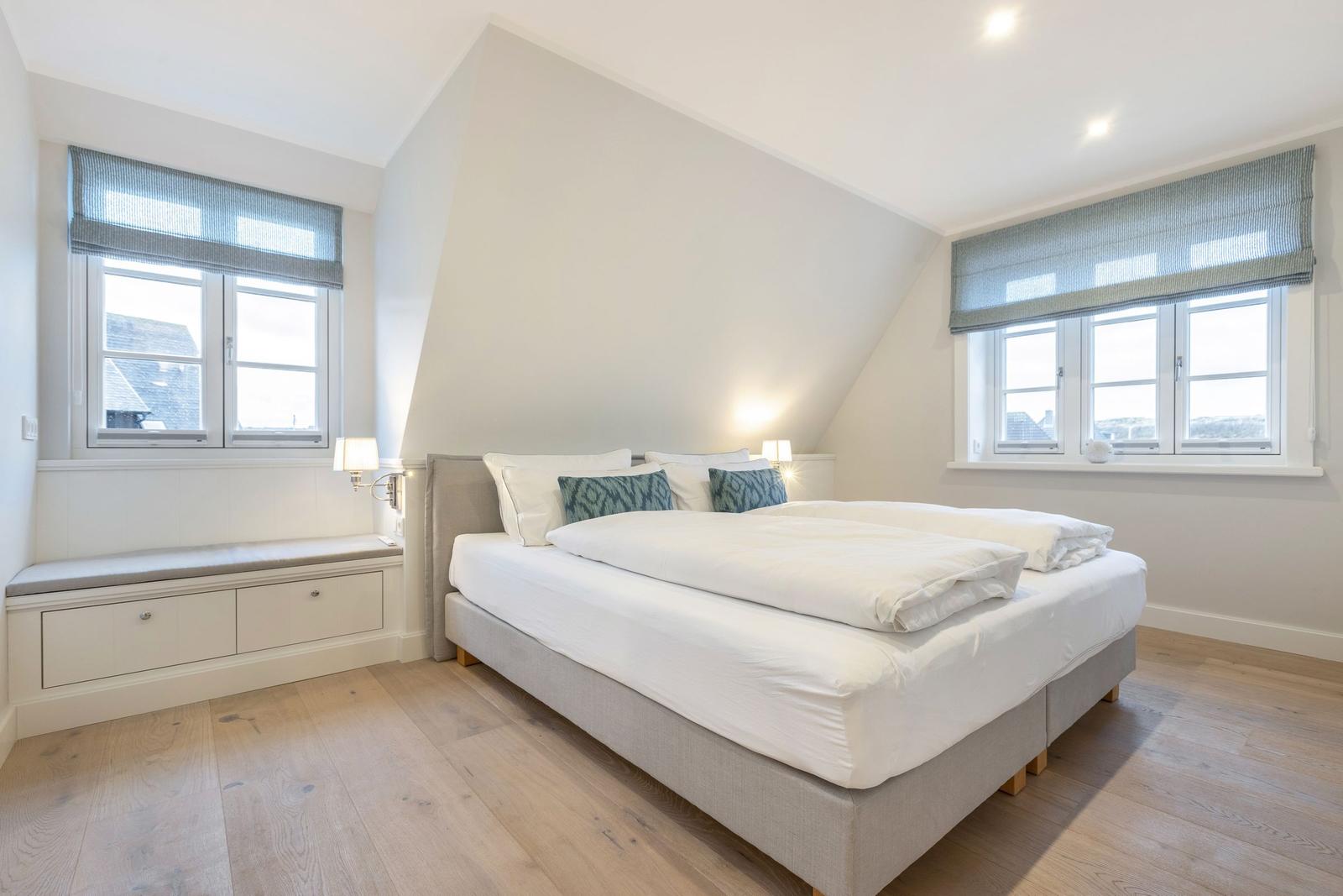 Schlafzimmer mit Doppelbett, zwei Fenstern und Holzboden.