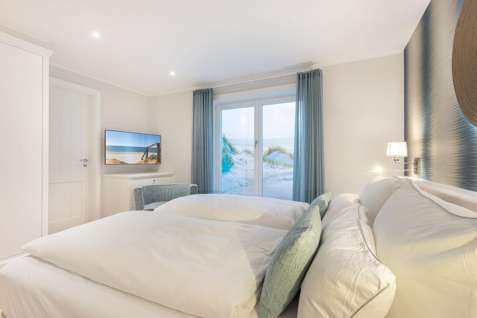 Schlafzimmer mit Blick auf Strand, TV und Bett mit Kissen.