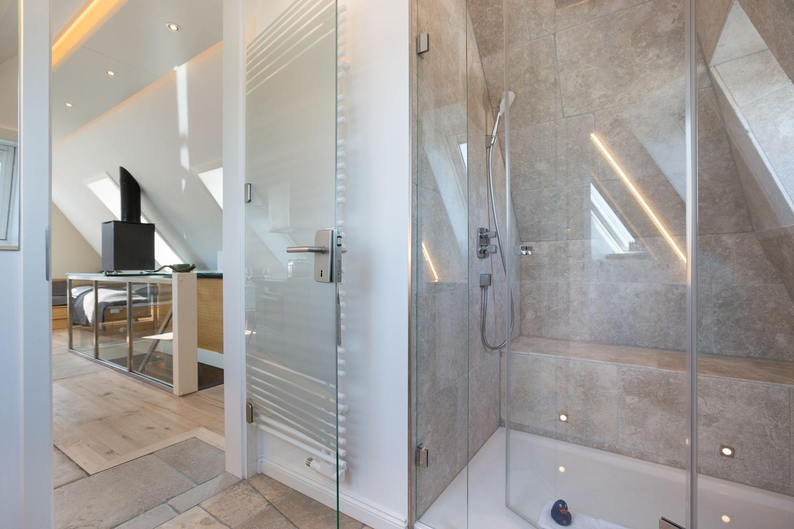 Dusche mit Glaswand und Duschbrause. Im Hintergrund Schlafzimmer mit Fenster.