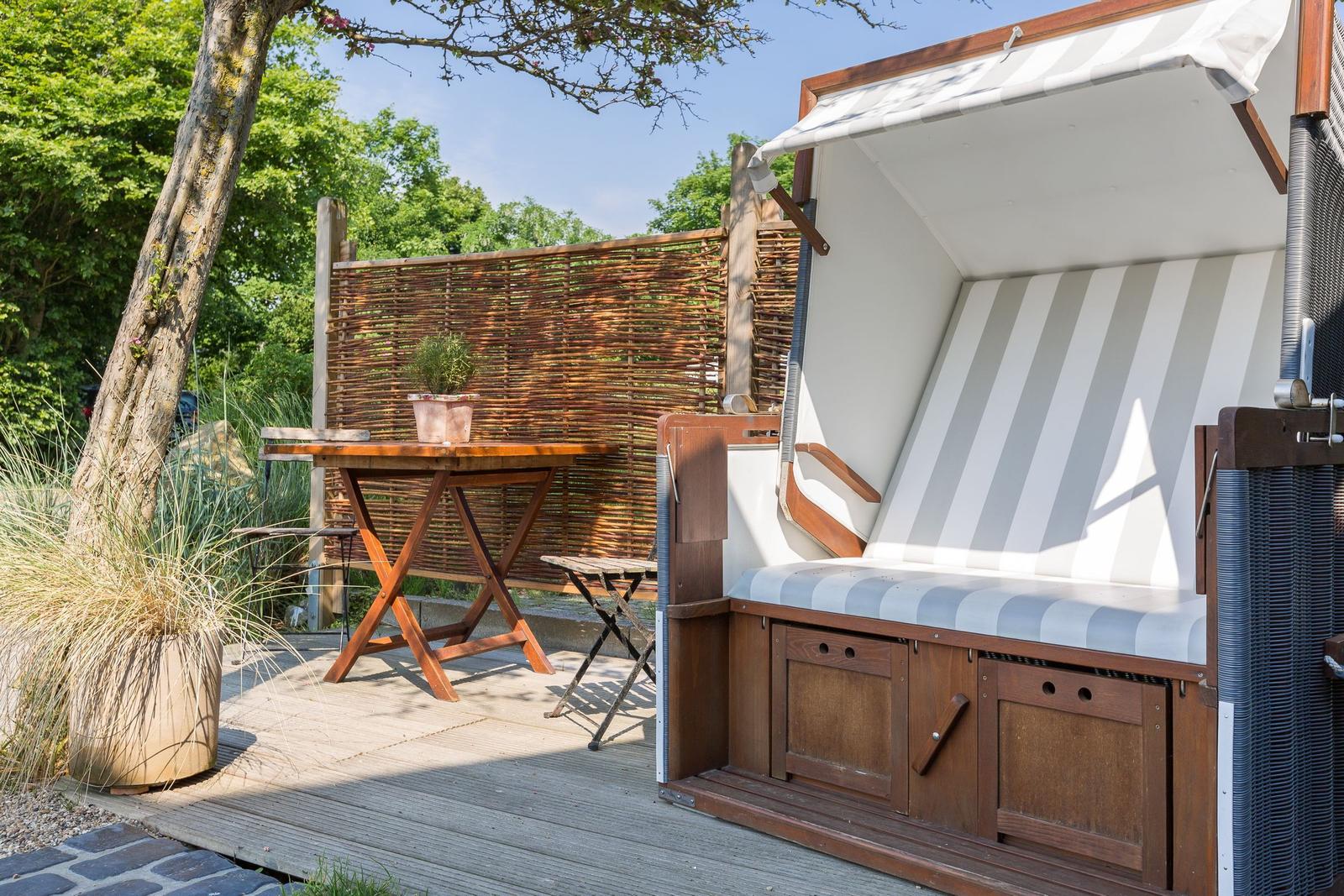 Hölzerner Strandkorb mit Streifenkissen auf Terrasse neben Tisch und Pflanzen.
