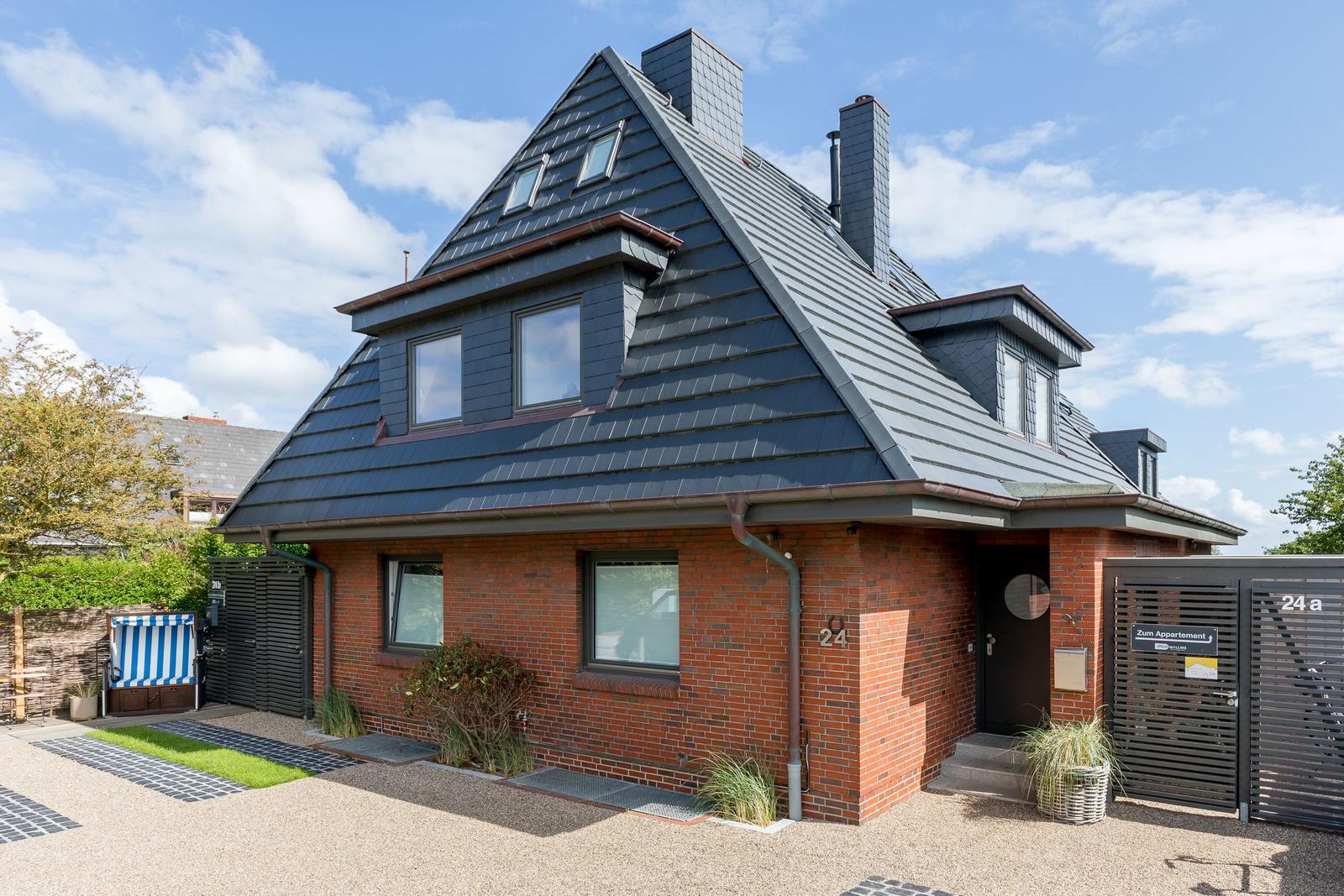 Ferienhaus mit rotem Backstein und dunklem Dach, Eingang mit Schild und Gartenbereich.