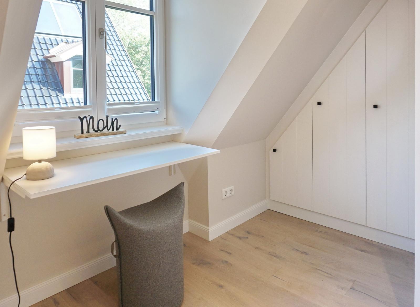 A work desk with chair and lamp under a window with sloped ceiling.