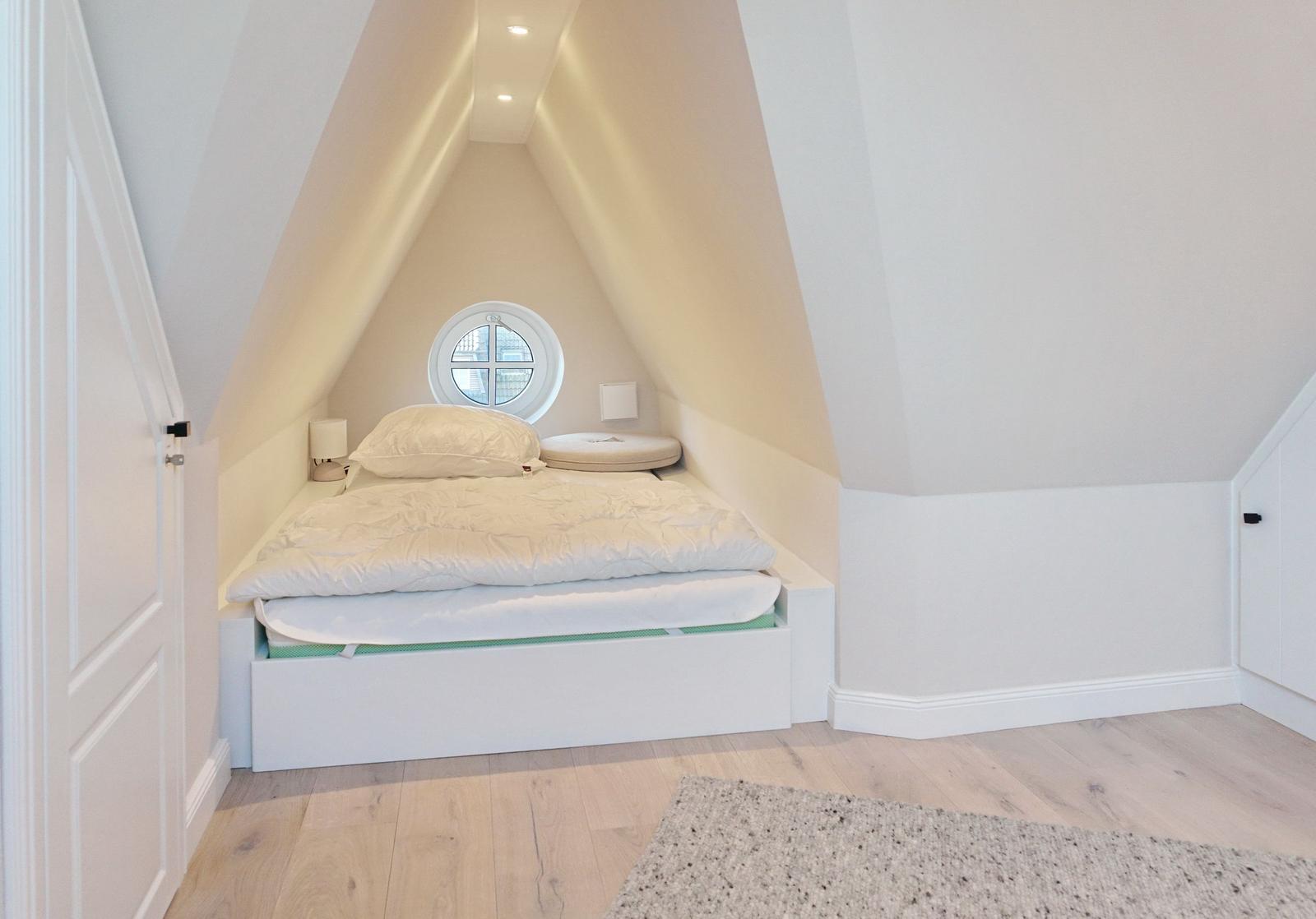 Bedroom with sloped ceiling, round window, and white bed.