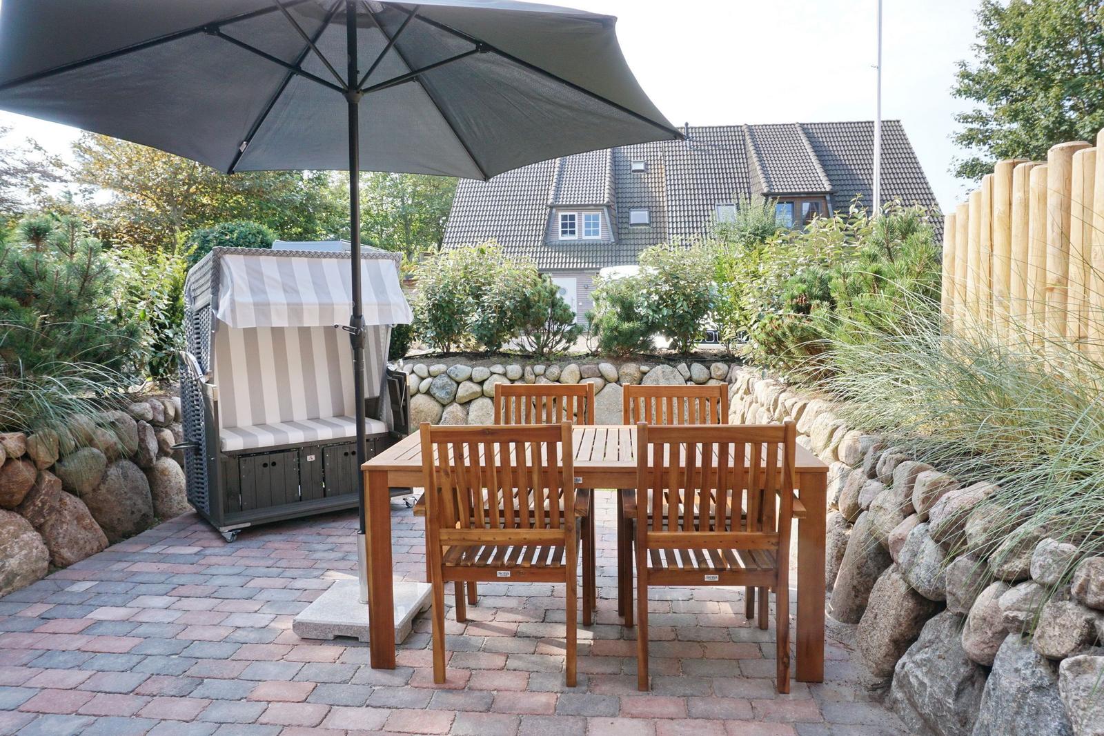 Terrace with table, chairs, and umbrella. Background: garden with stone wall and house.