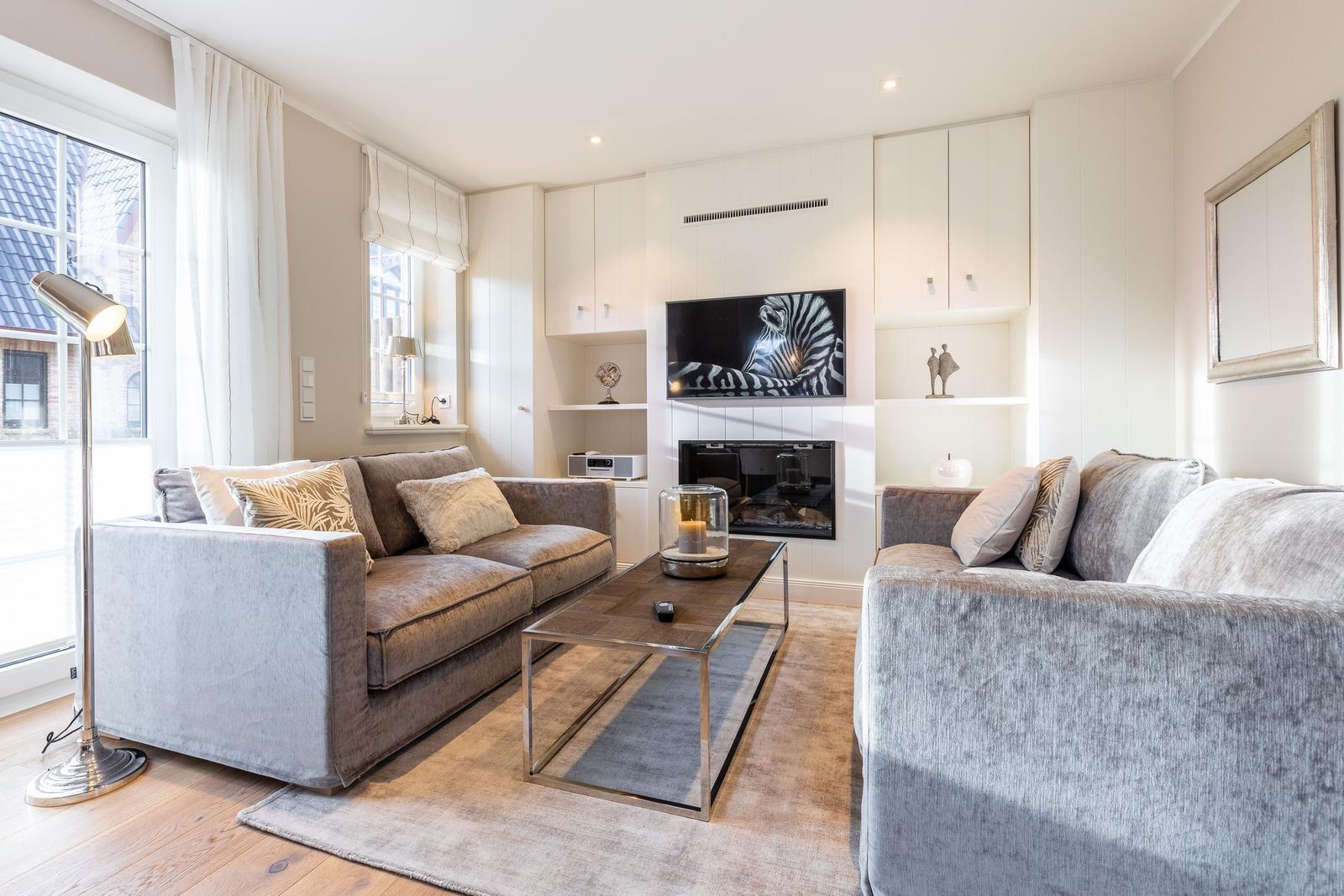 Living room with two sofas, TV, and fireplace. Wooden floor and large windows.