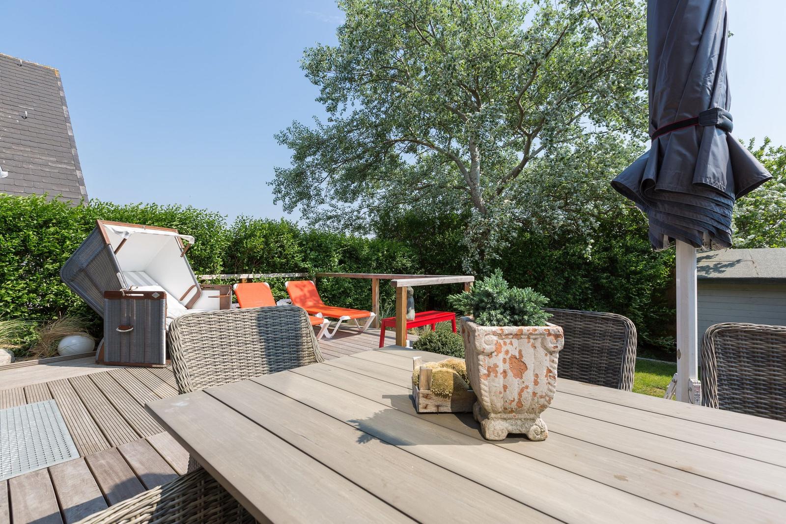 Terrasse mit Tisch, Stühlen und Liegestühlen. Hintergrund: Baum und Hecke.