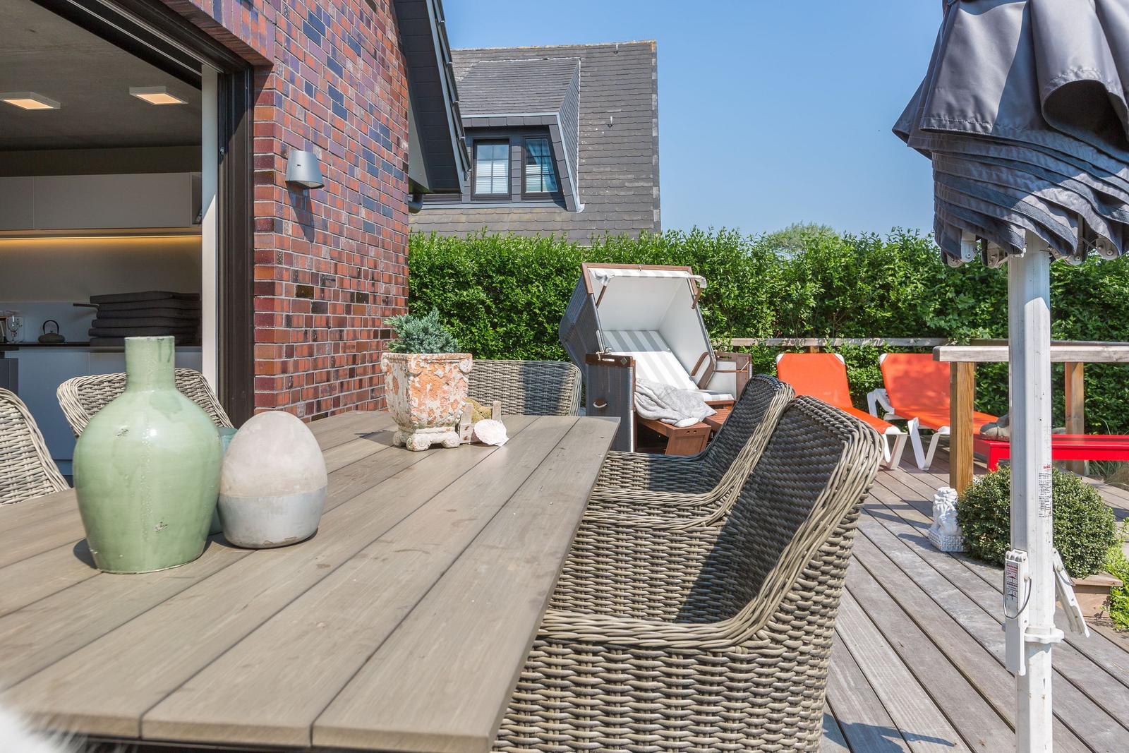 Terrasse mit Tisch, Stühlen und Sonnenschirm. Hintergrund: grüne Hecke und Haus.