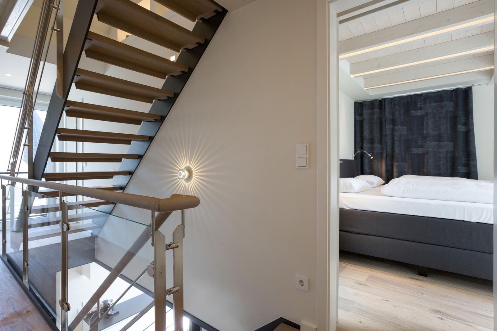Treppenhaus mit Holzstufen und Glasgeländer. Blick in Schlafzimmer mit Bett und Vorhang.