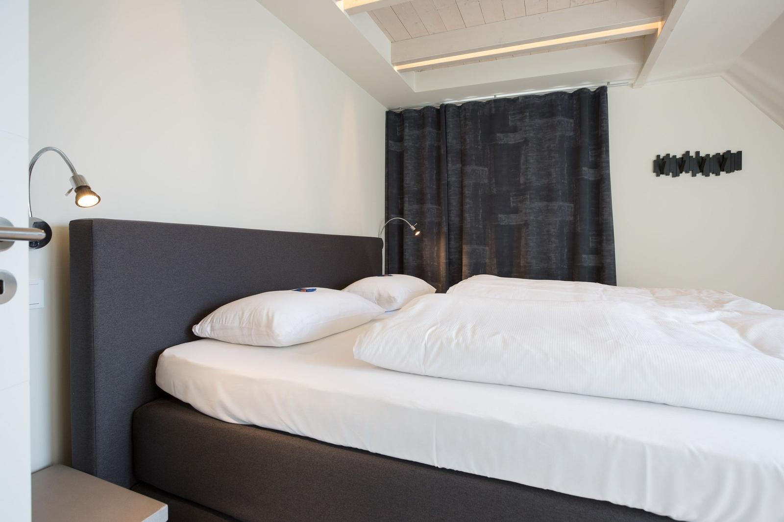Schlafzimmer mit Bett, weißen Laken und dunklen Vorhängen.