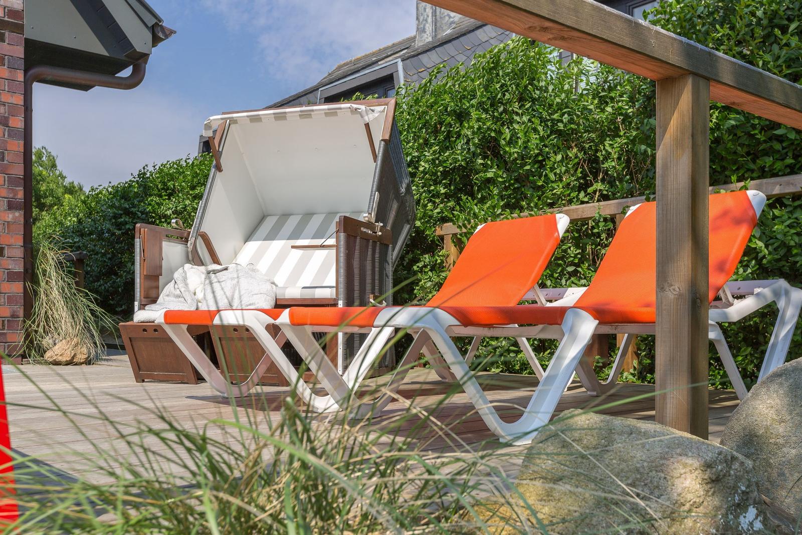 Terrasse mit orangefarbenen Liegestühlen und Sonnenschirm. Holzgeländer und Grünflächen im Hintergrund.