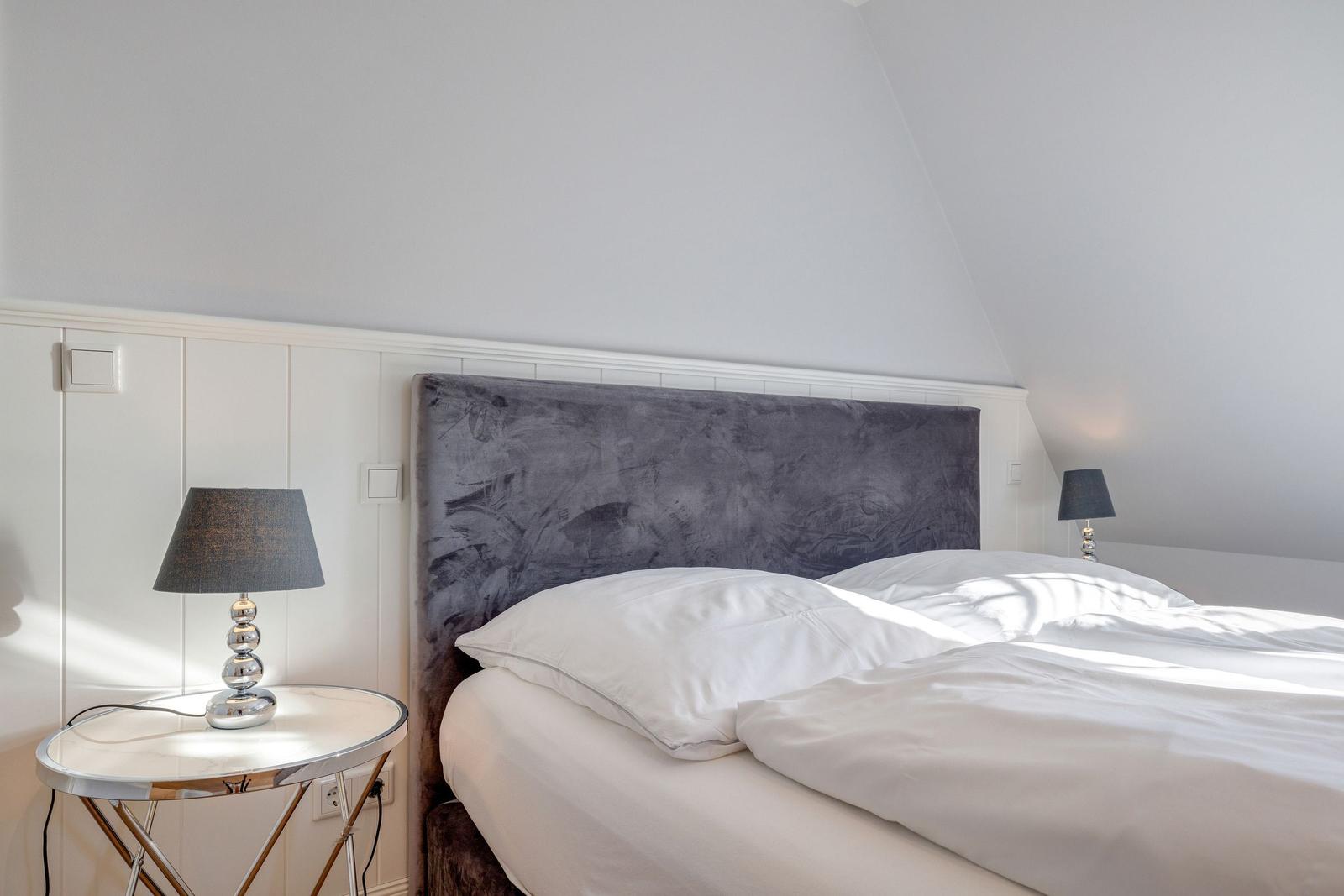 Schlafzimmer mit Bett, grauem Kopfteil und zwei Nachttischen mit Lampen.