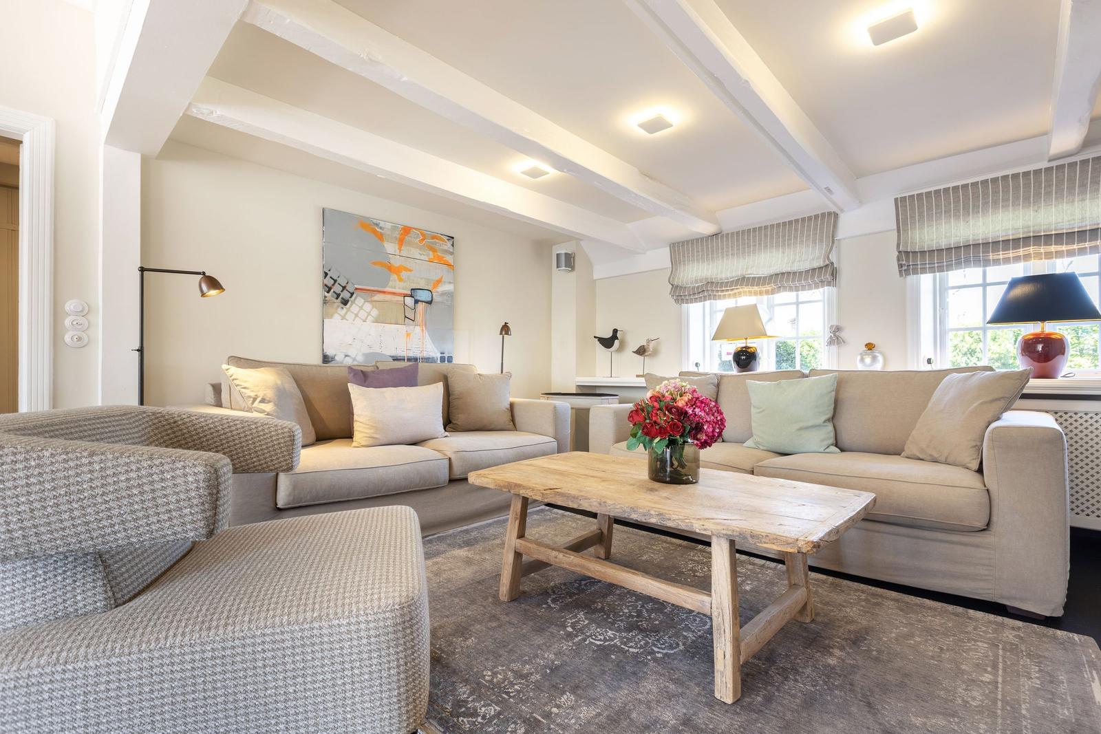 Wohnzimmer mit zwei Sofas, Holztisch und Blumen. Deckenbalken und Fenster mit Jalousien.