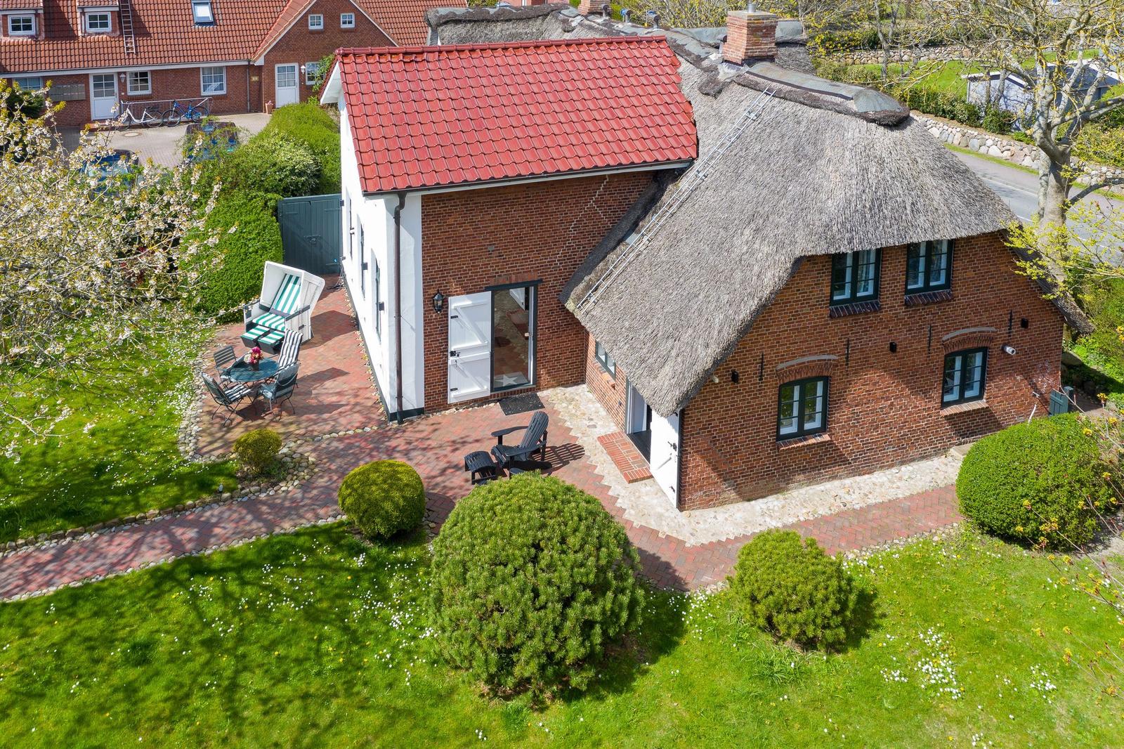 Rotes Dach, strohgedeckter Teil, Terrasse mit Gartenmöbeln und Rasenfläche.