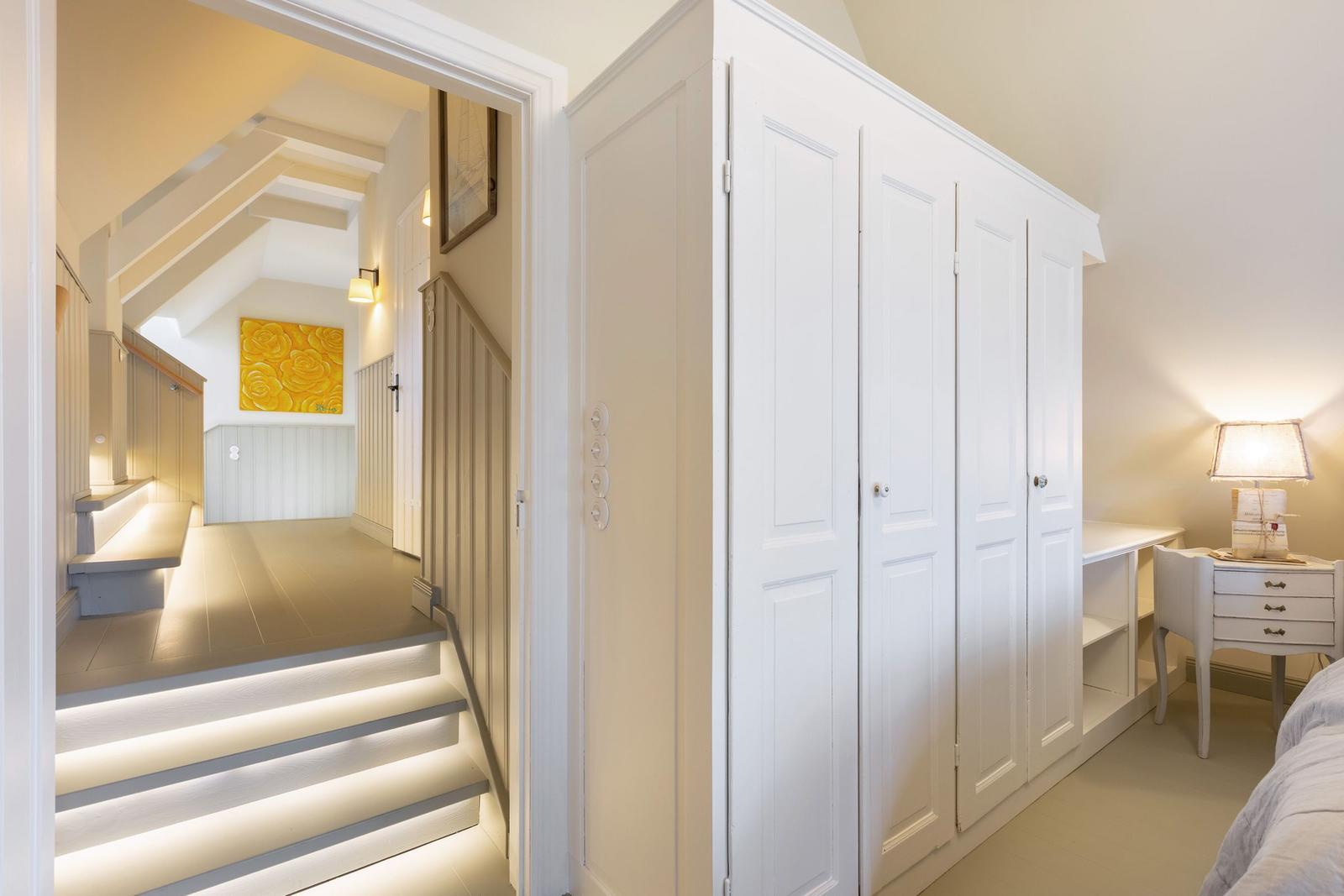 Schlafzimmer mit weißem Kleiderschrank und Nachttisch. Treppenhaus mit beleuchteten Stufen und gelbem Wandbild.