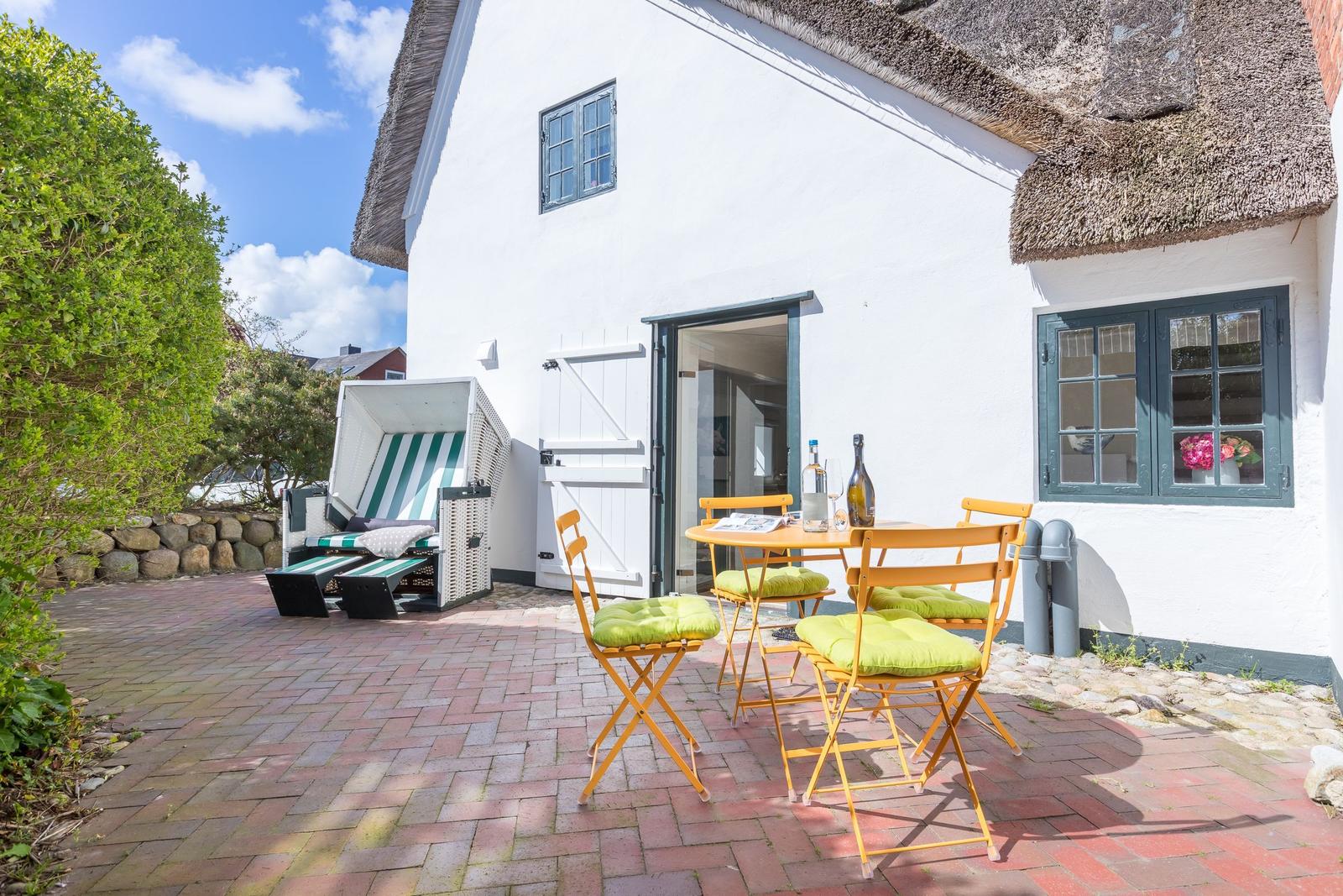 House with terrace, table, chairs, and beach chair. Thatched roof and white walls.