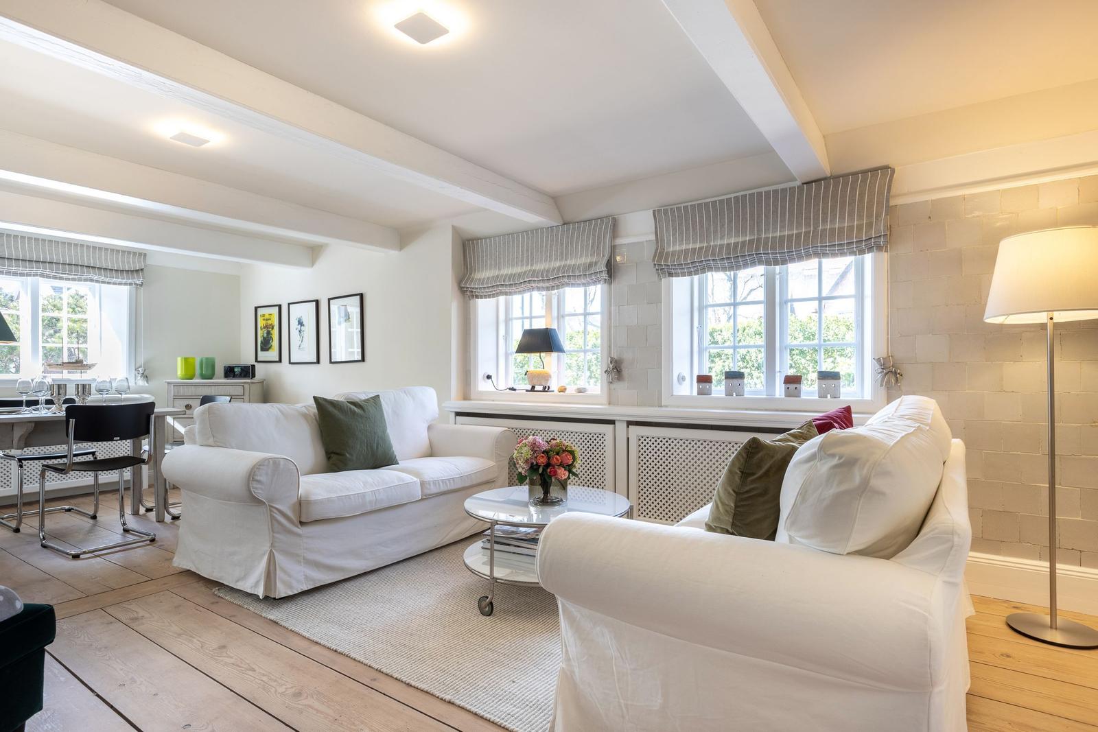 Living and dining area with white sofas, wooden floor, and windows with blinds.