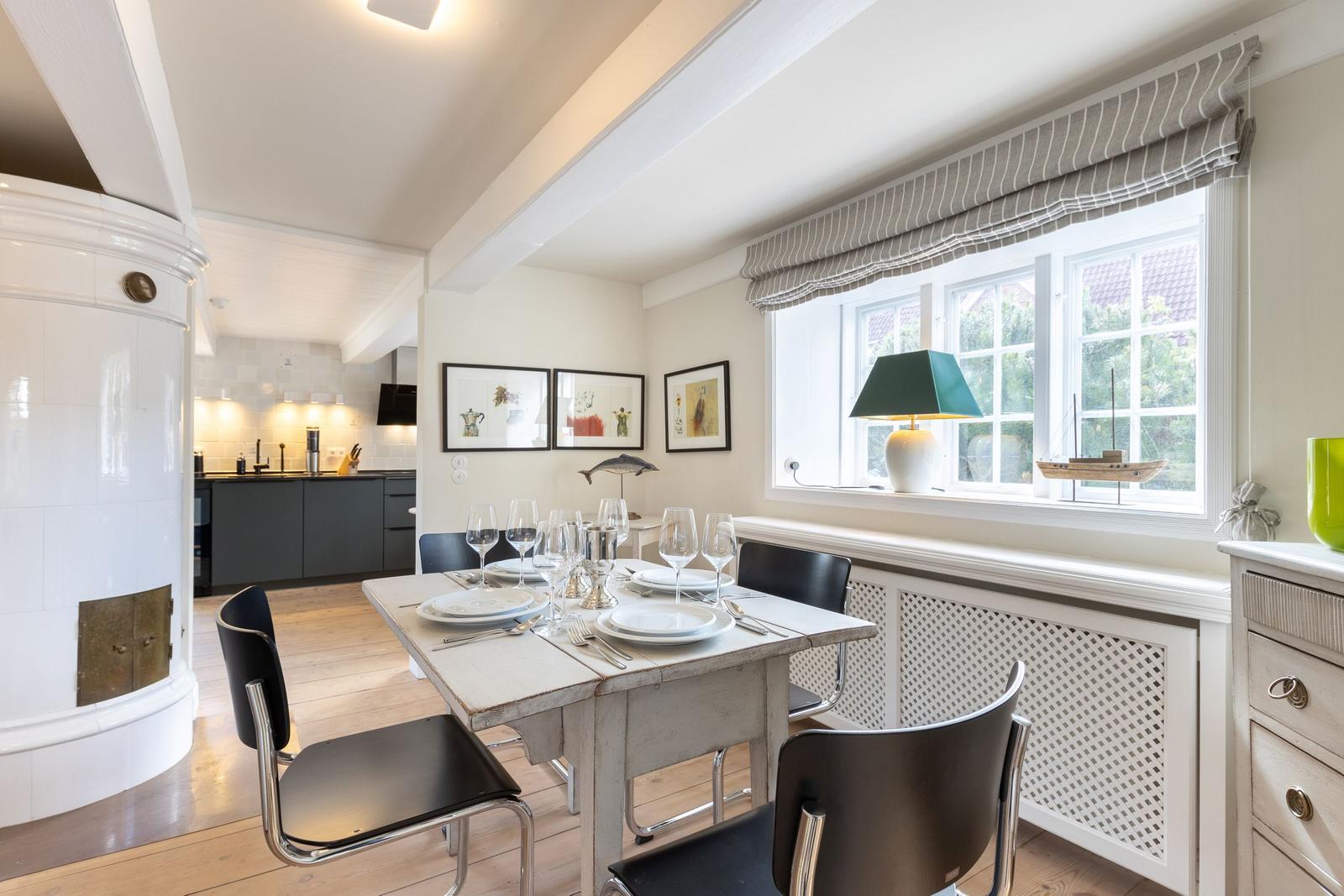 Dining room with table, chairs, and window. Kitchen in background. White walls and wooden floor.