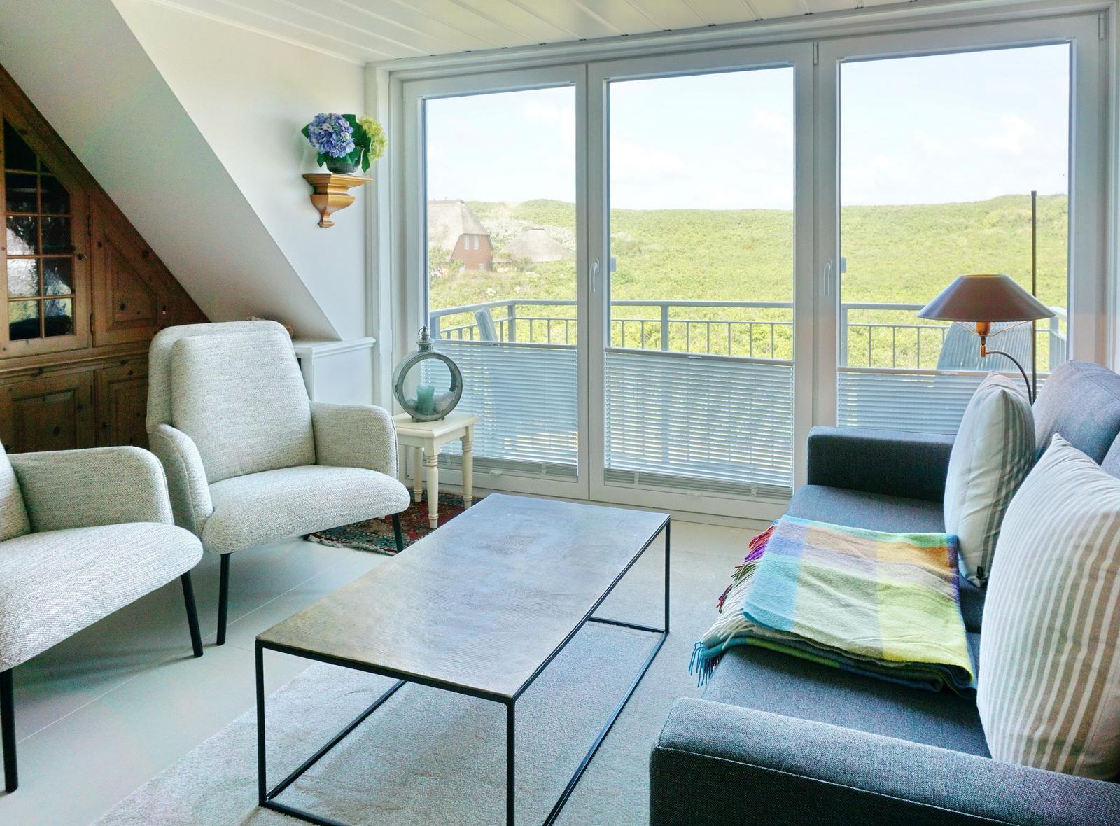 Wohnzimmer mit Sofa, Sessel und Tisch. Balkon mit Blick auf grüne Hügel.