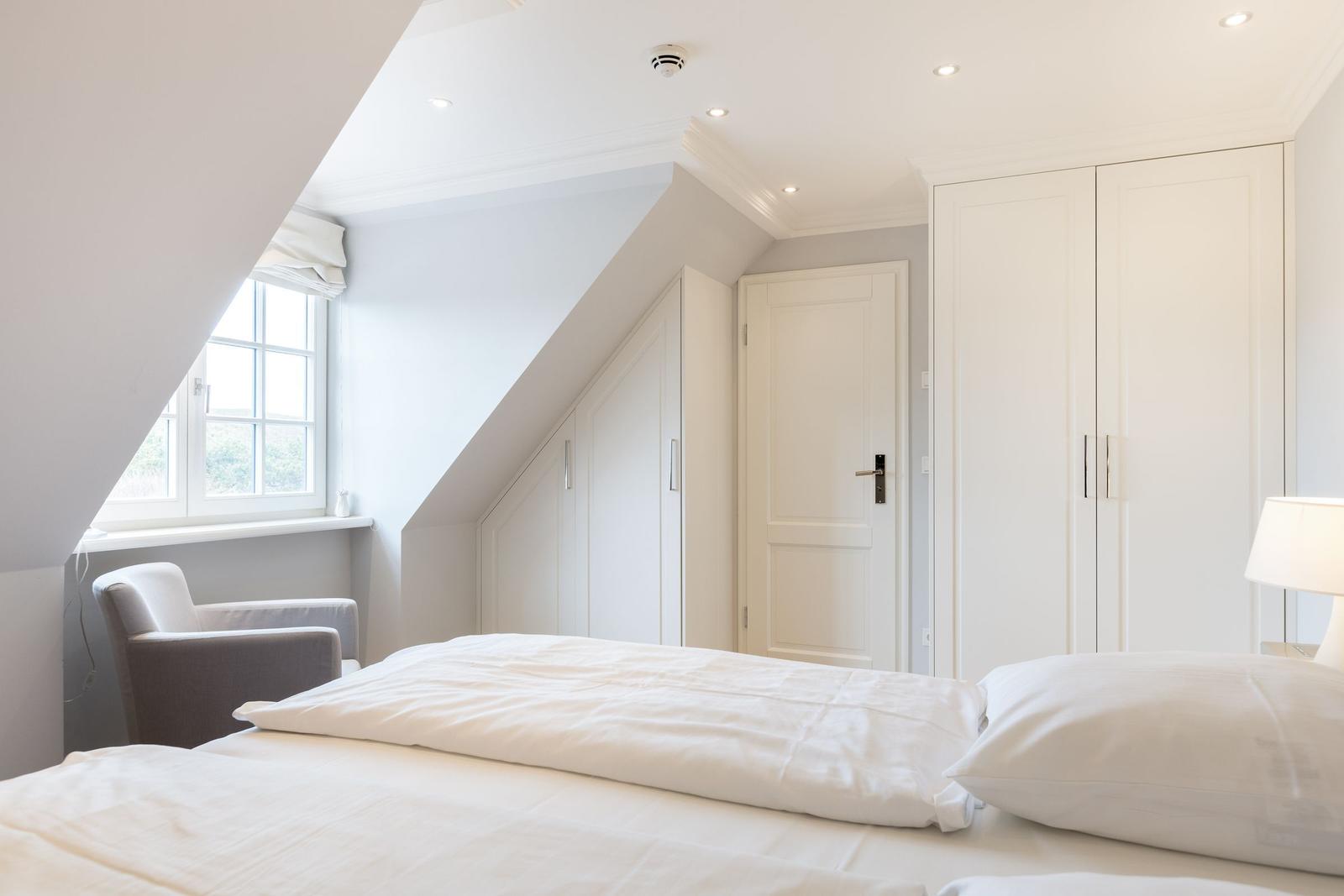 Schlafzimmer mit Dachschrägen, weißem Bett, Kleiderschrank und Fenster.