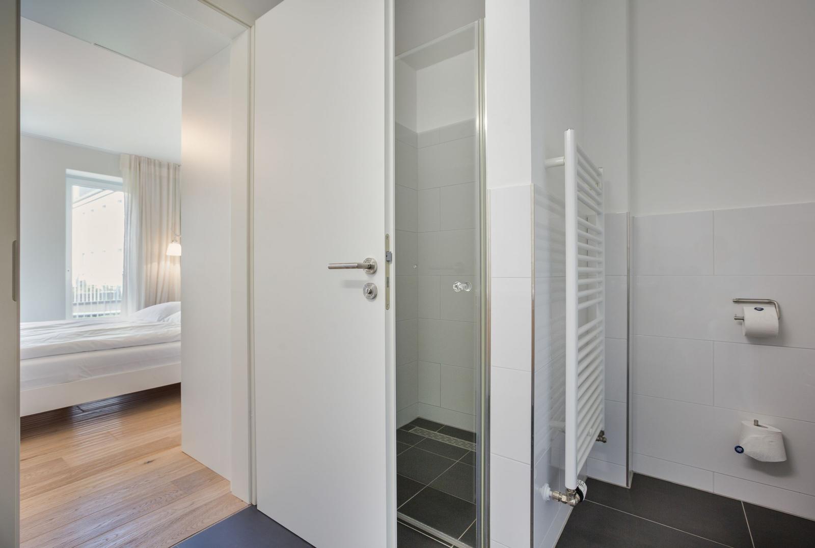 Badezimmer mit Dusche und Heizkörper. Durchgang in Schlafzimmer mit Bett und Fenster.