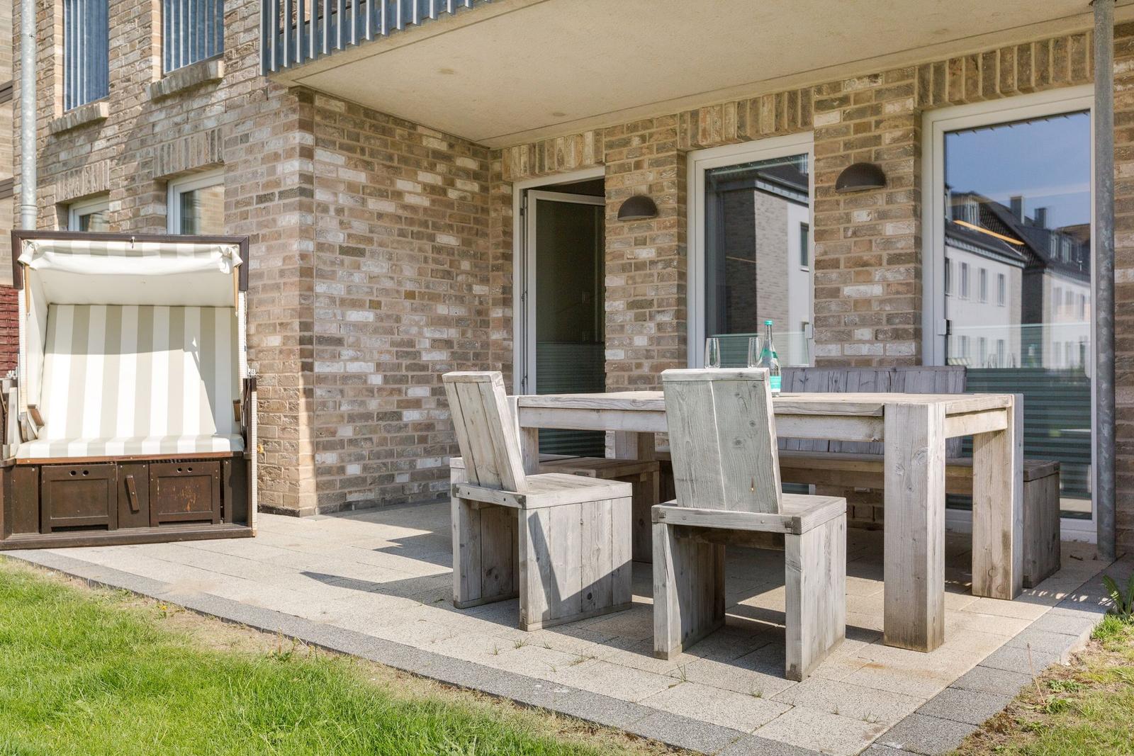 Terrasse mit Holztisch, Stühlen und Strandkorb. Hintergrund: Backsteinhaus mit Fenstern.