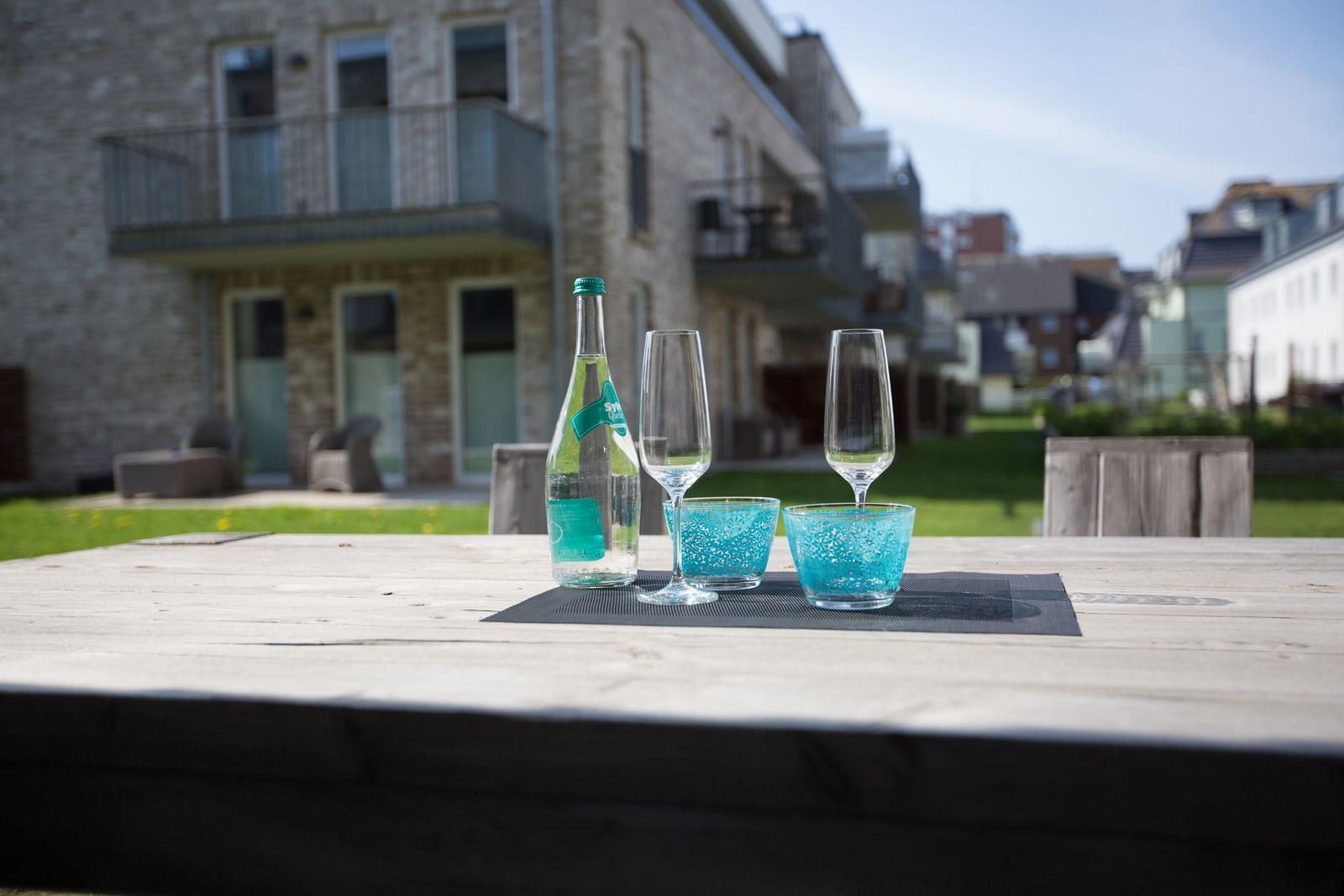 Außenbereich mit Holztisch, Flasche und Gläsern. Hintergrund: Steinhaus mit Balkonen.