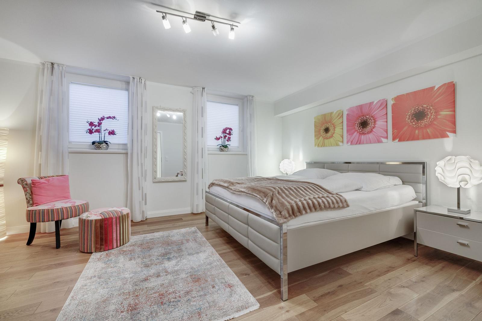 Schlafzimmer mit Bett, zwei Fenstern, Spiegel und Blumenbildern an der Wand.