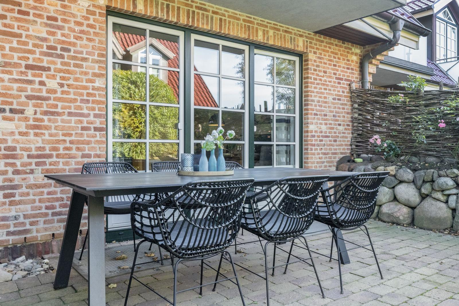 Terrace with table and chairs in front of brick house with windows.