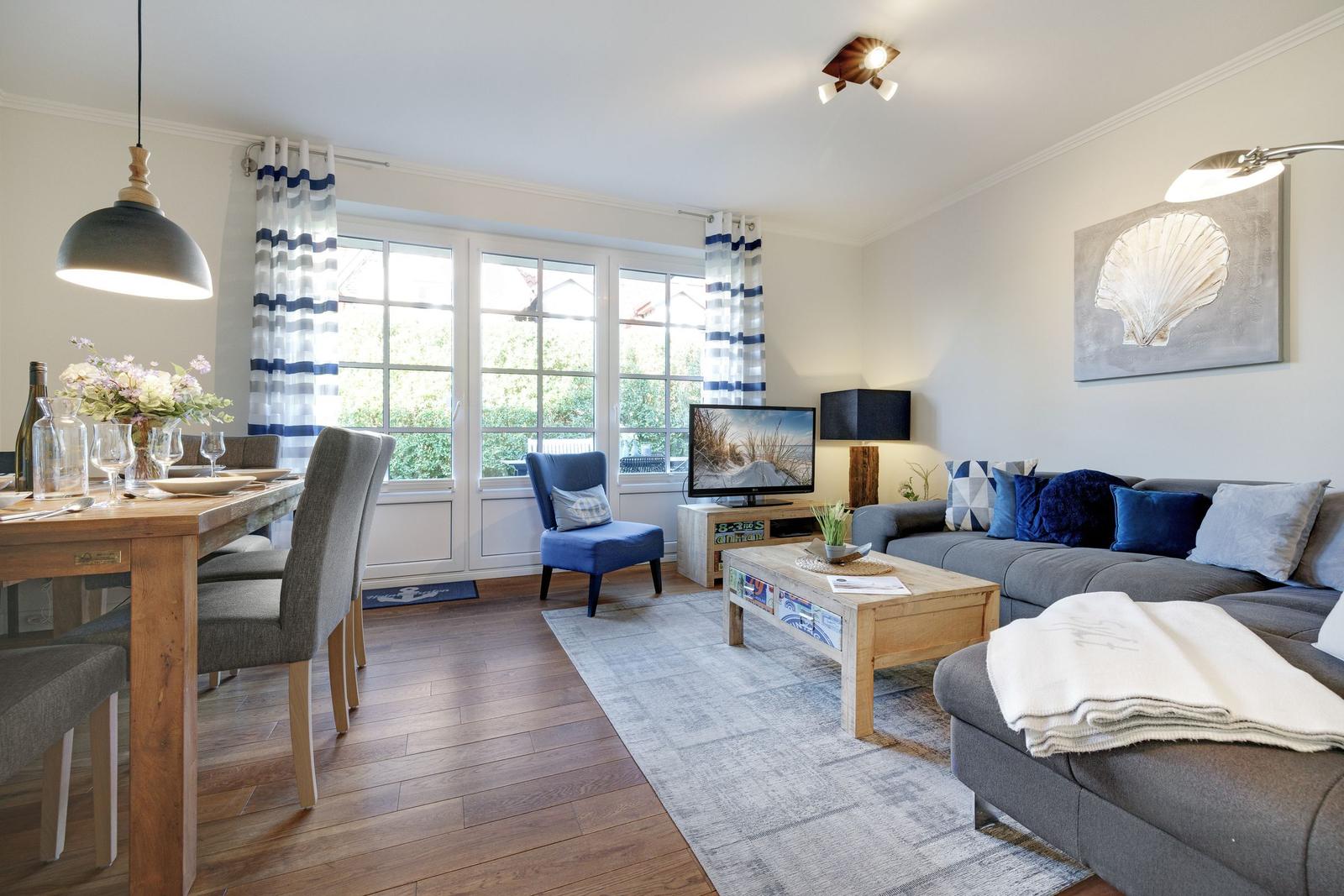Living and dining area with sofa, coffee table, and TV. Wooden floor and large windows.