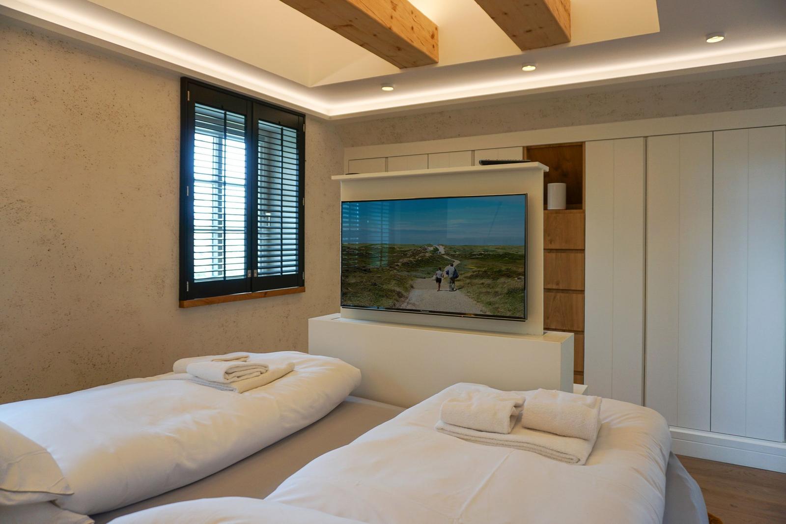 Two beds with white linens, TV, and window with blinds.