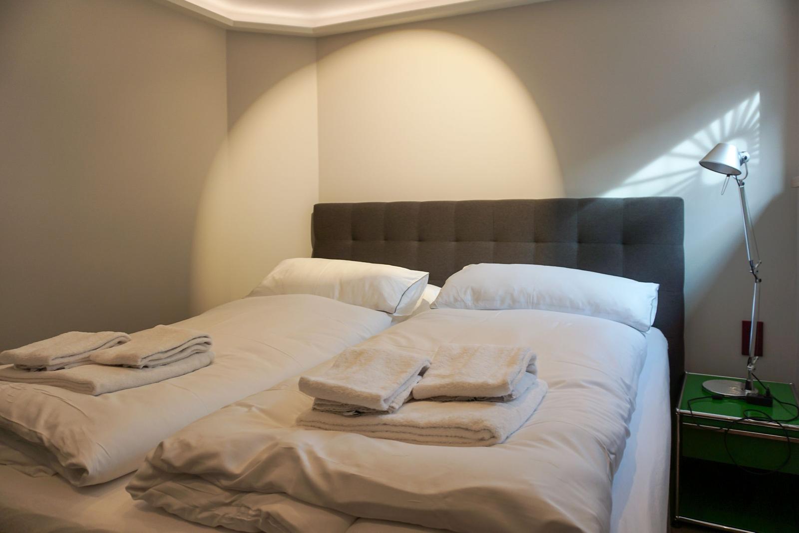 Double bed with white bedding and folded towels. Green nightstand with floor lamp.