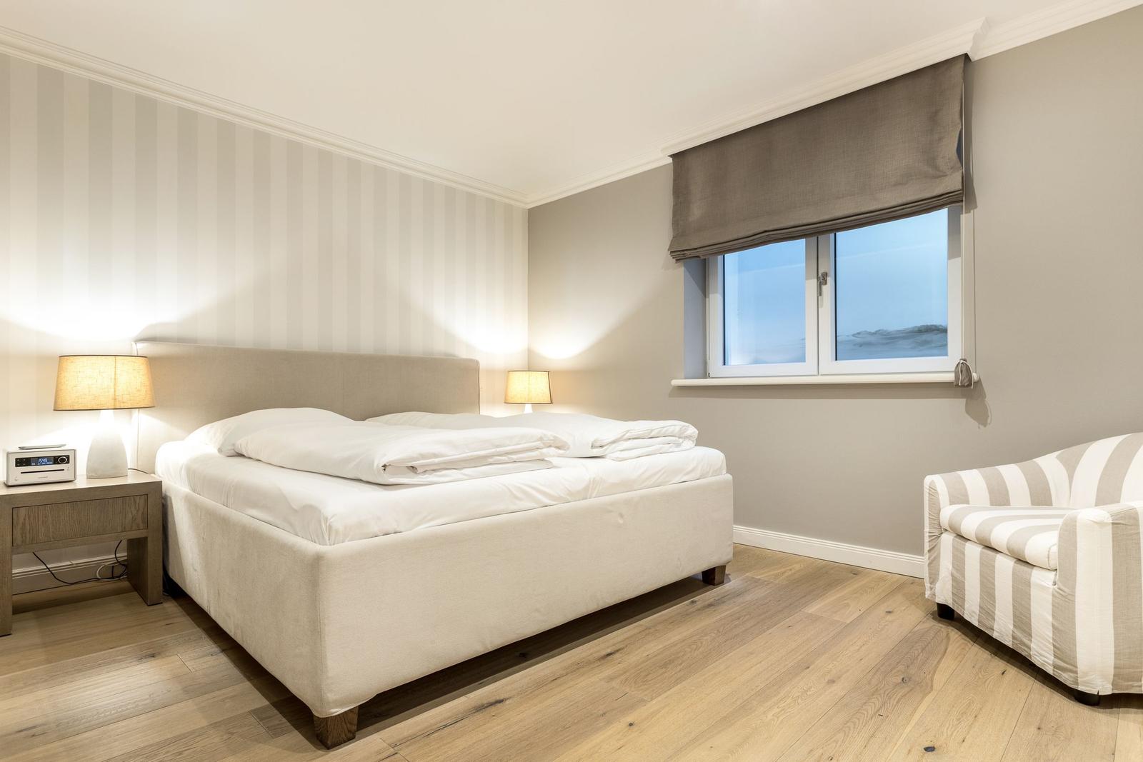 Bedroom with bed, nightstand, lamp, and armchair. Wooden floor and window with roller blind.