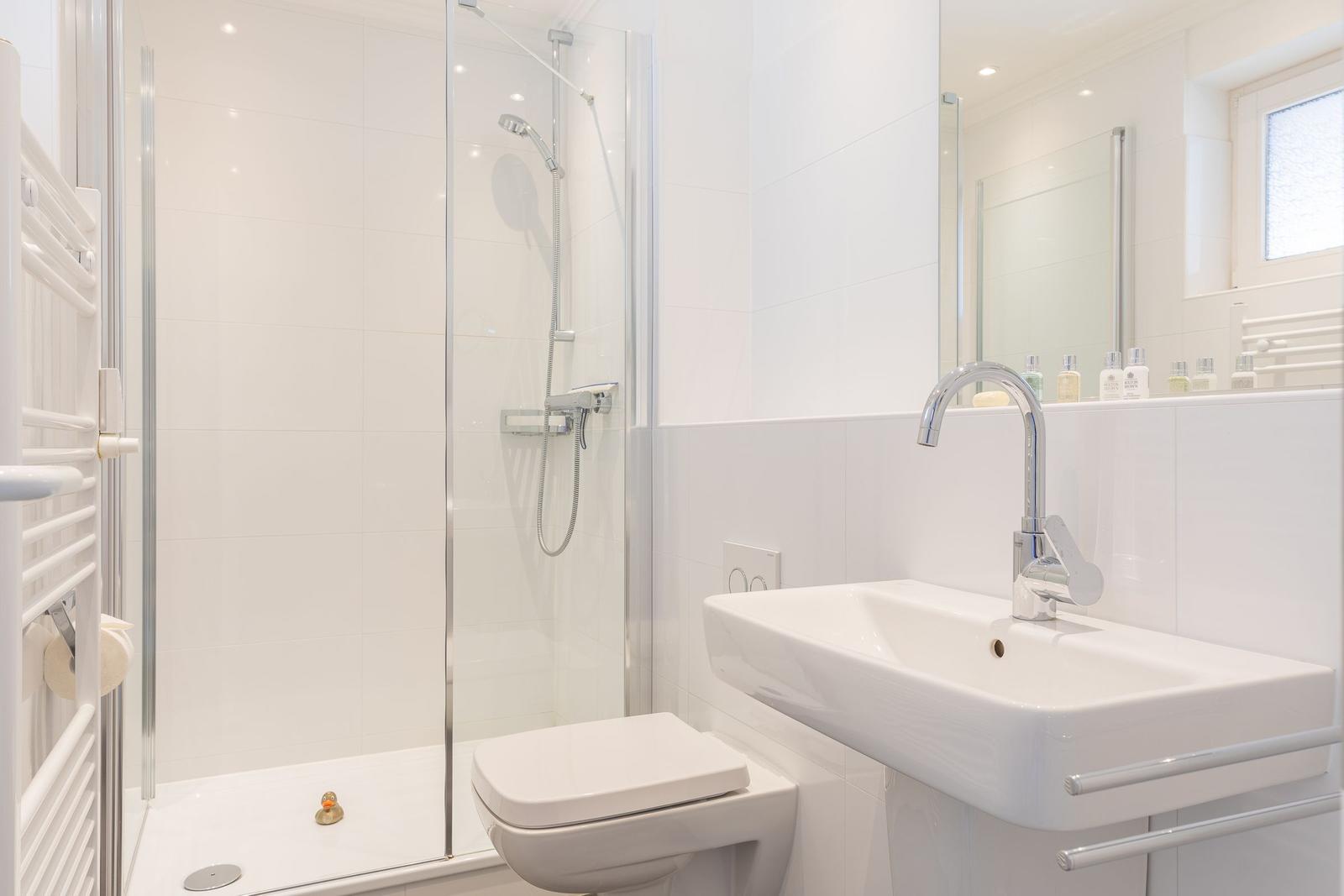 Badezimmer mit Dusche, Waschbecken und Toilette. Weiße Fliesen und Spiegel.