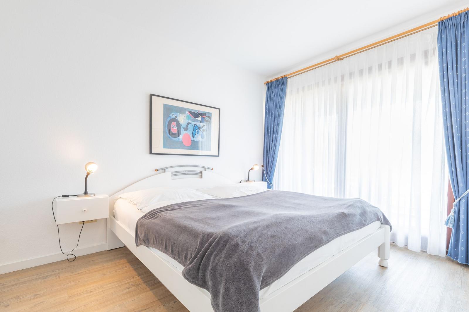 Schlafzimmer mit weißem Bett, grauer Decke und blauen Vorhängen.