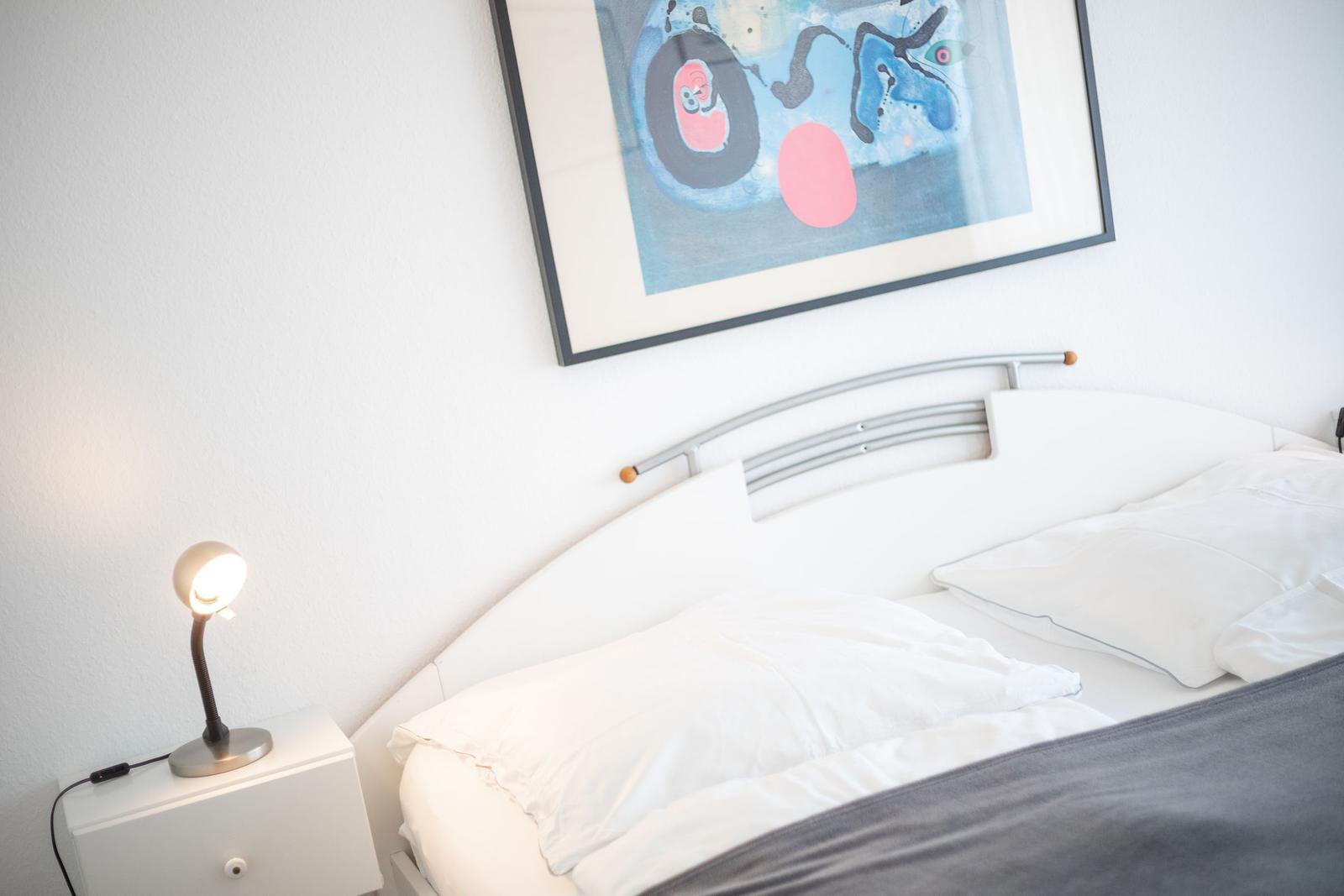 Ein Bett mit weißen Kissen und einer grauen Bettdecke. An der Wand hängt ein abstraktes Bild.