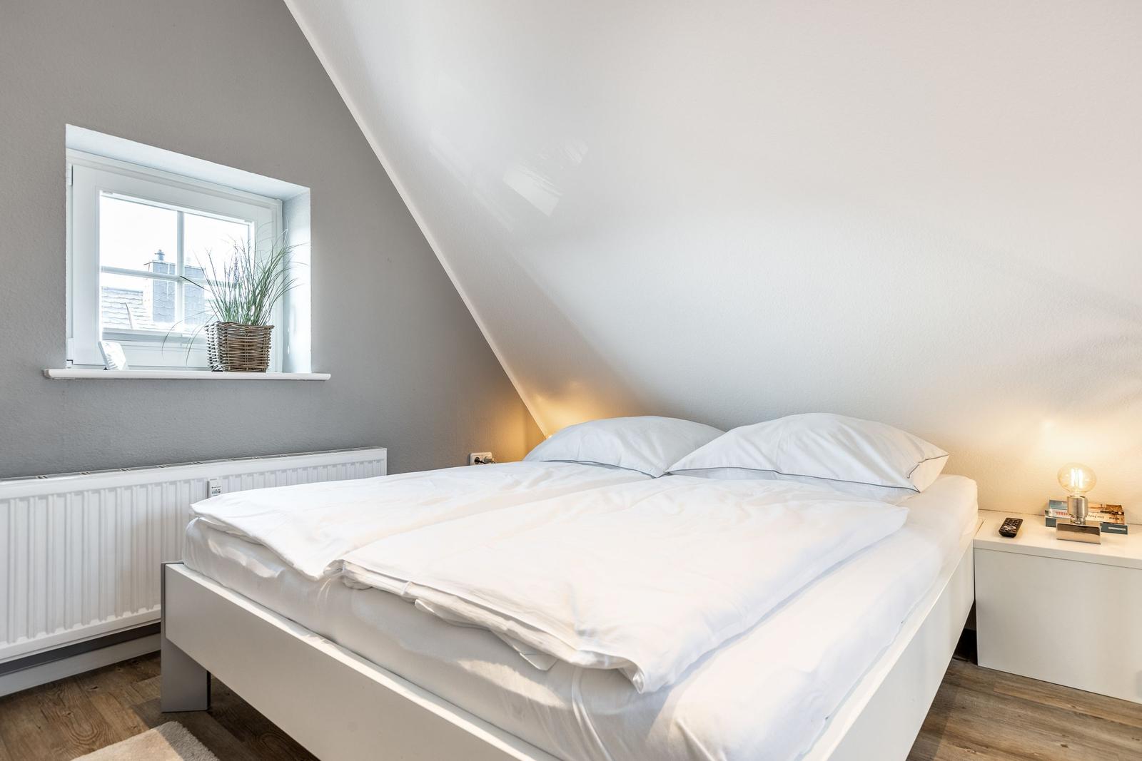 Bedroom with sloped ceiling, white bed, and window with plant.