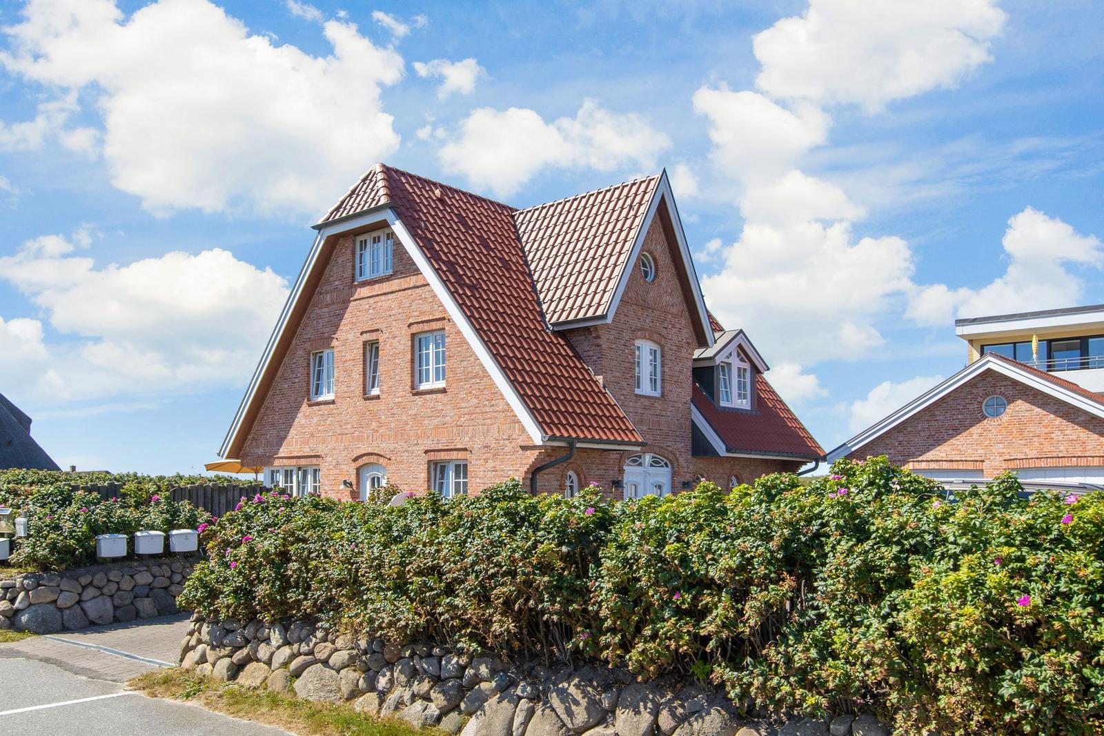 Rotes Ziegeldach, weiße Fensterläden, grüne Hecke mit rosa Blüten.